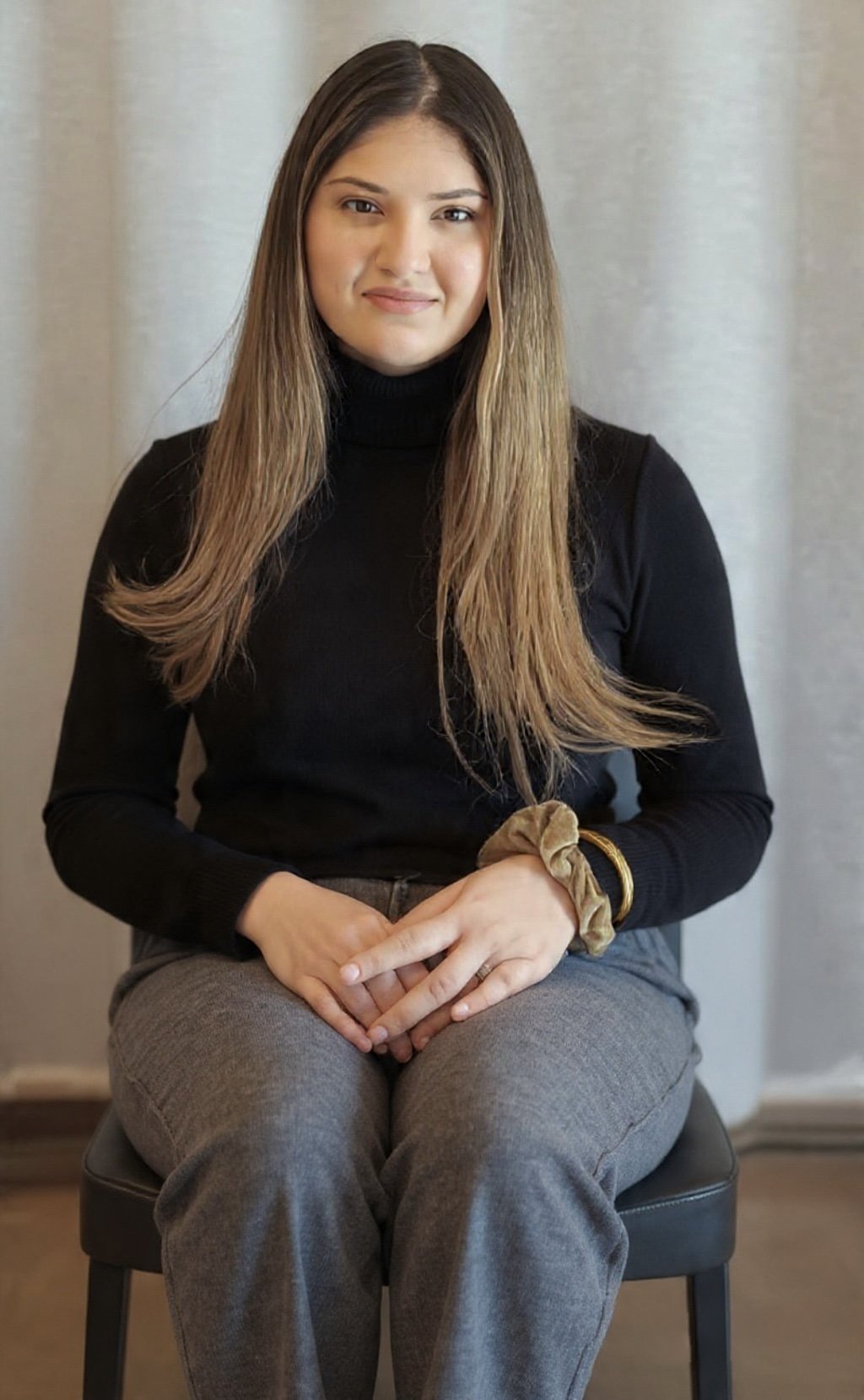 A young woman with long, light brown hair sitting on a black chair. She is wearing a black turtleneck sweater, gray pants, and a tan scrunchie on her wrist. She has a subtle smile and is looking at the camera against a neutral background.