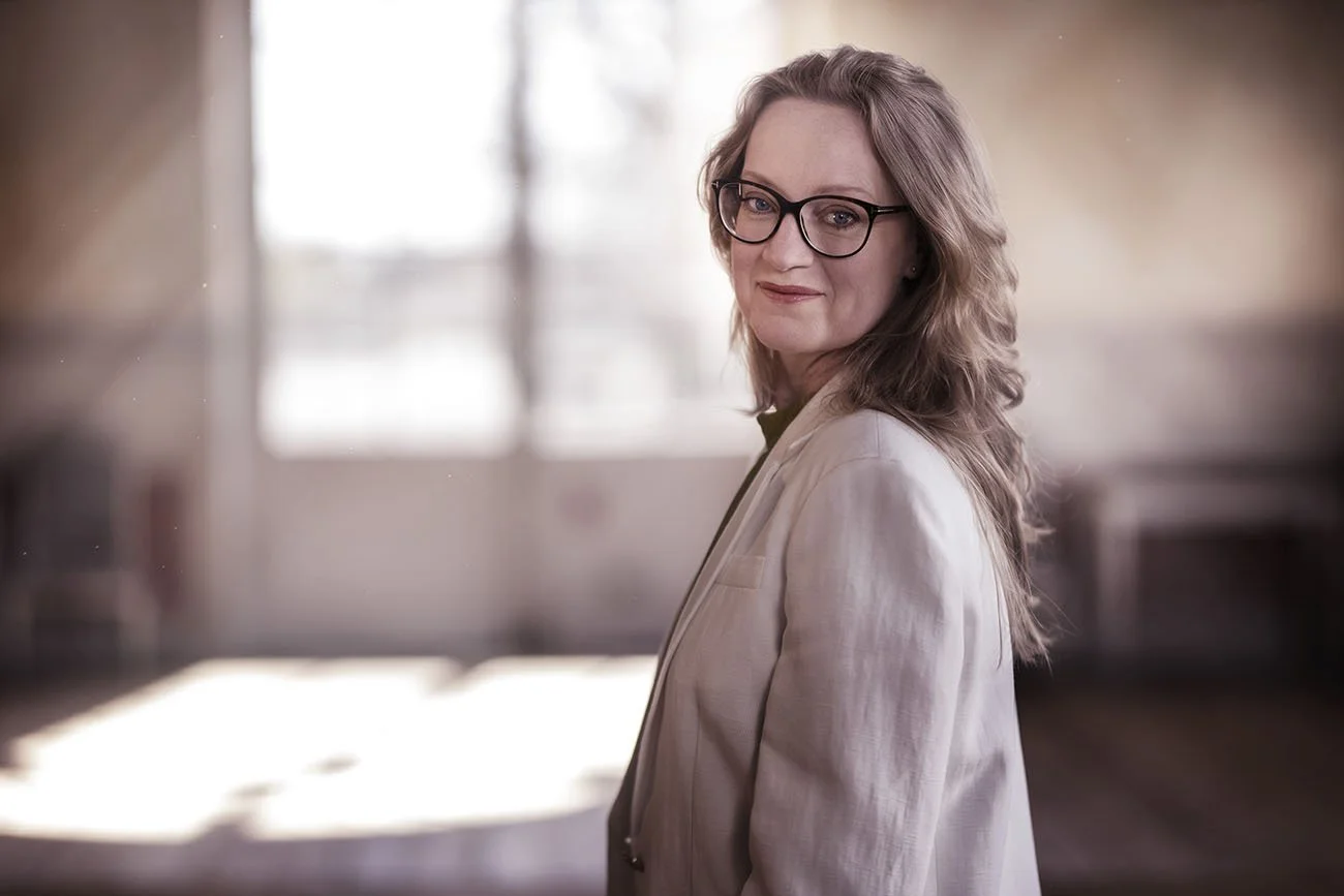 En kvinna med glasögon och ljus jacka, som tittar mot kameran, i ett inomhusmiljö med ljusa fönster i bakgrunden.