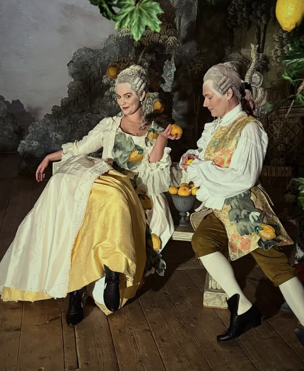 Två personer i historiska kostymer poserande med citroner i en konstnärlig bakgrund. Damen sitter elegant medan herren står lutad med koppar. De är omgivna av gröna växter och ett dekorativt bakgrundssmålning.