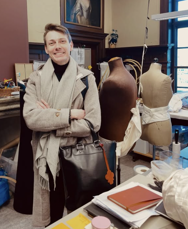 En man står i ett rum med två mannekänger, ett av dem är en tennfigur och det andra är en tygmodell. Mannen är glad och har på sig en beige kappa och en grå scarf. Det finns olika kontors- eller konstmaterial på bordet framför honom.