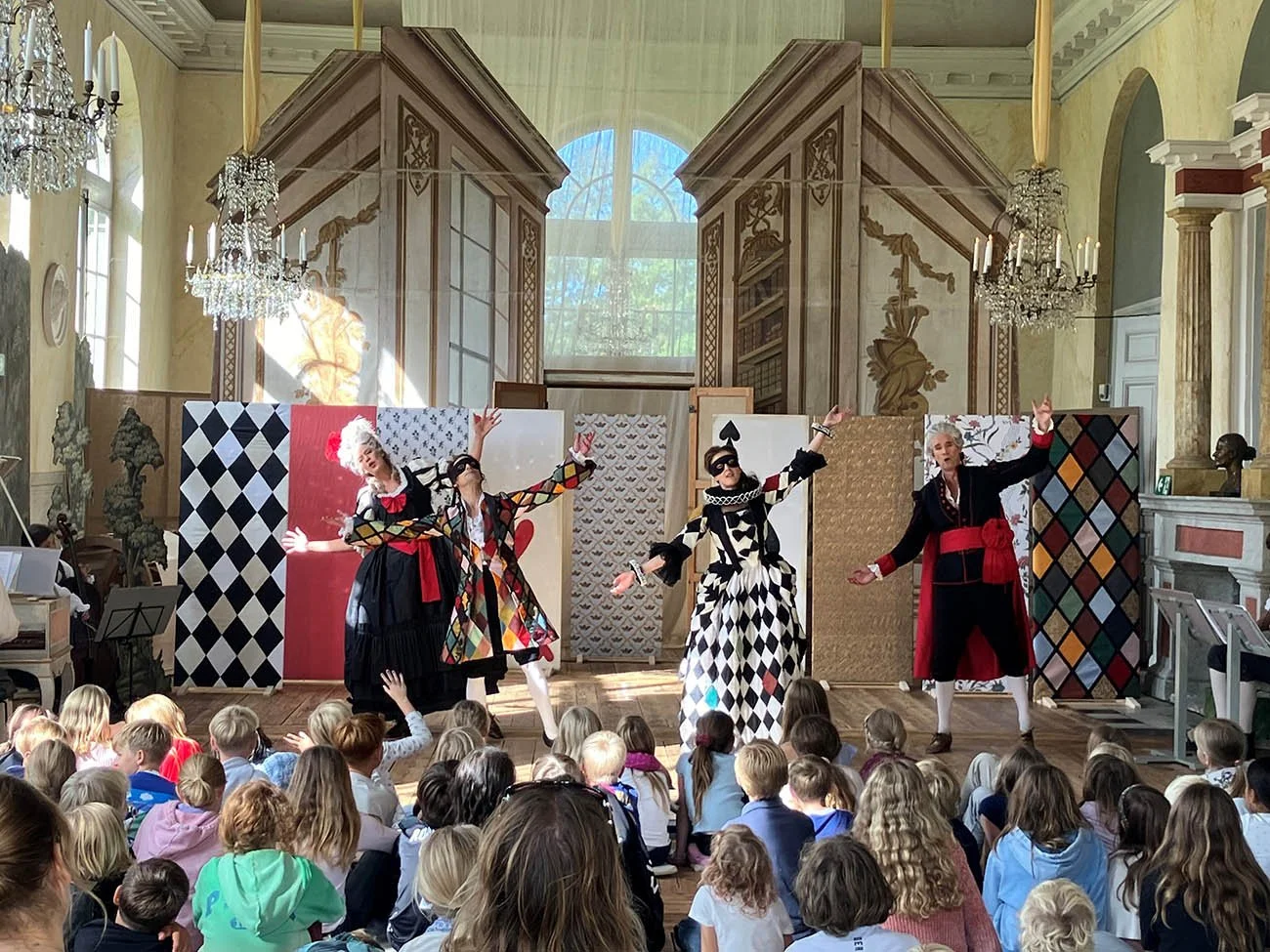 Fyra personer i kostym framför en publik av barn och vuxna, som spelar teater på en scen i ett elegant rum med stora fönster, kristallkronor och fantasifull bakgrund.