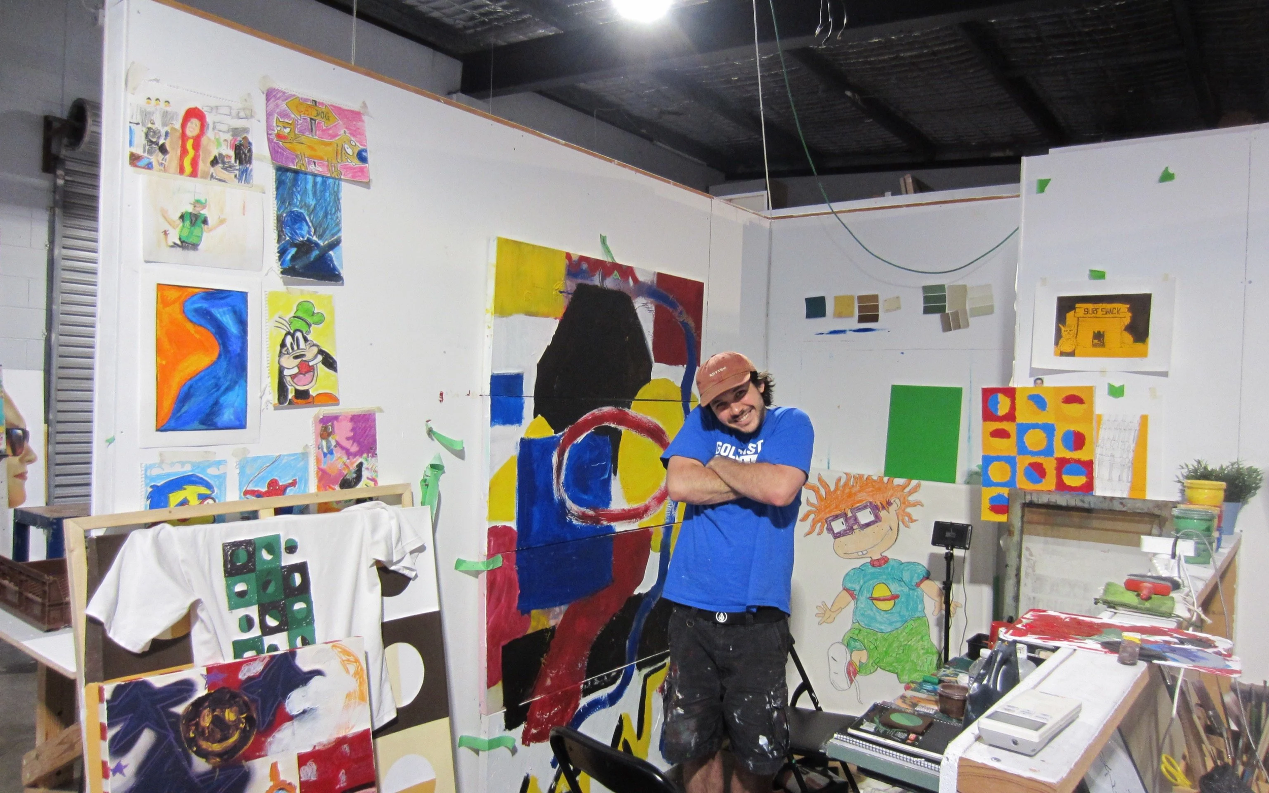 A smiling man in a blue shirt and cap stands with arms crossed in an art studio filled with colorful paintings and art supplies. The walls are covered with various artwork, including cartoon characters and abstract designs.