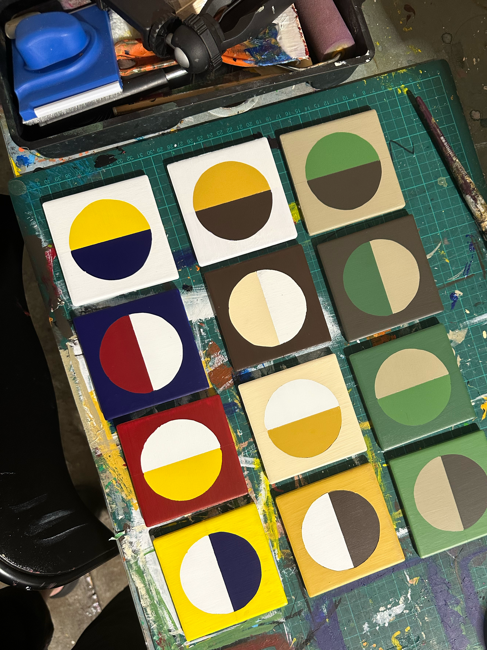 A table with several square art canvases painted with half-circle designs in various colors, arranged in rows. The background is cluttered with painting supplies, including brushes, paints, and a cutting tool.