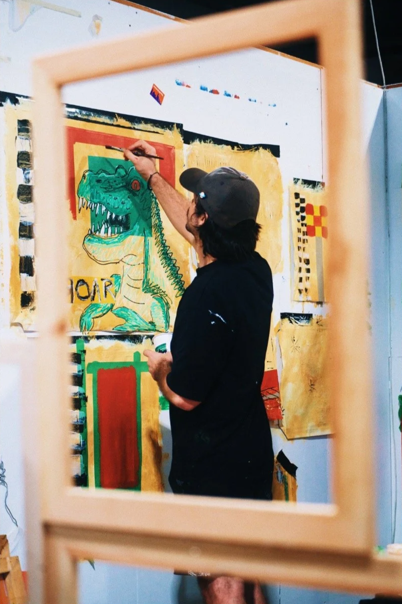 An artist is painting a cartoonish green dinosaur with sharp teeth on a large paper or canvas, seen through a wooden frame.