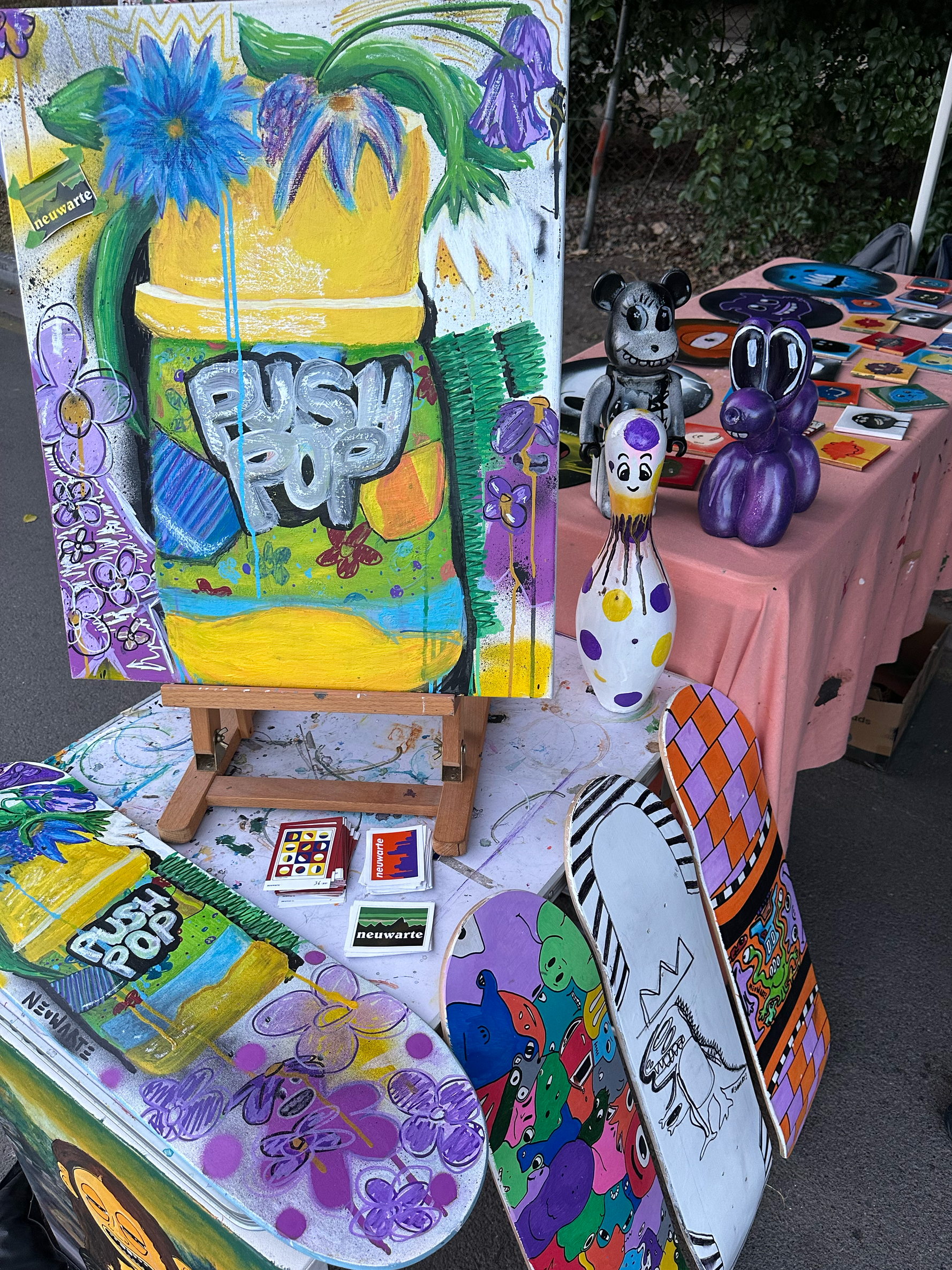 An art booth displays colorful paintings, small sculptures, and skateboards with designs. The paintings feature vibrant flowers, abstract patterns, and the words 'Push Pop.' Sculptures include a gray mouse with black eyes, a purple bunny, and a polka