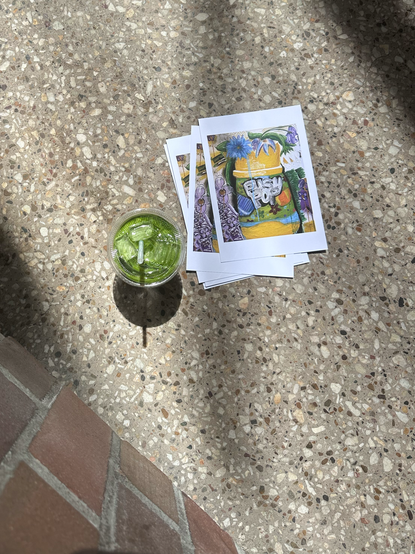 A cup of green slush with a straw next to a stack of colorful printed photographs of a painted summer scene with flowers, a beach, and the words "Push Pop".