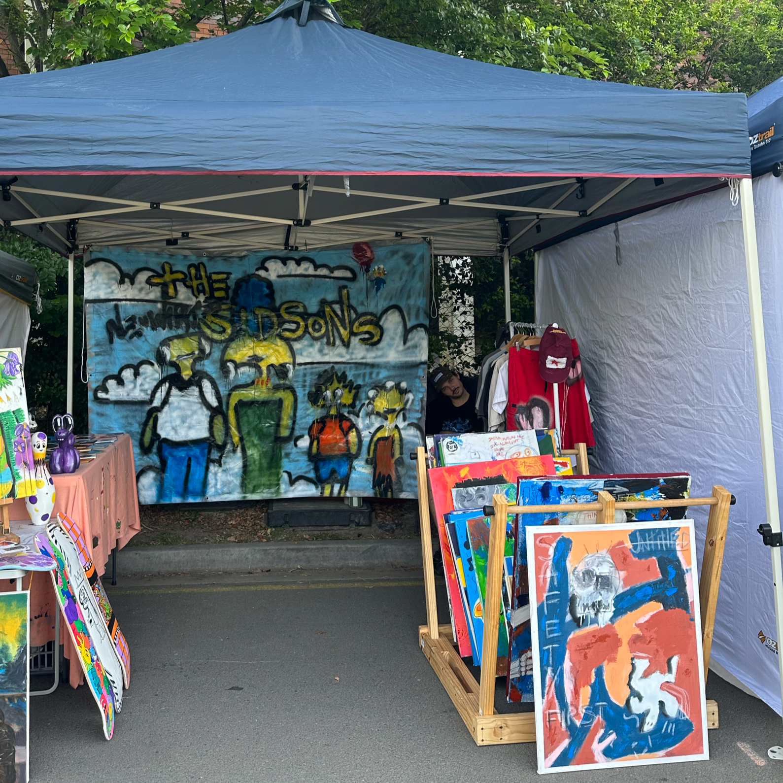 An outdoor art vendor booth under a grey canopy displaying colorful paintings, including a large banner with abstract human figures and stormy clouds, and various smaller paintings and art pieces on display stands and racks.