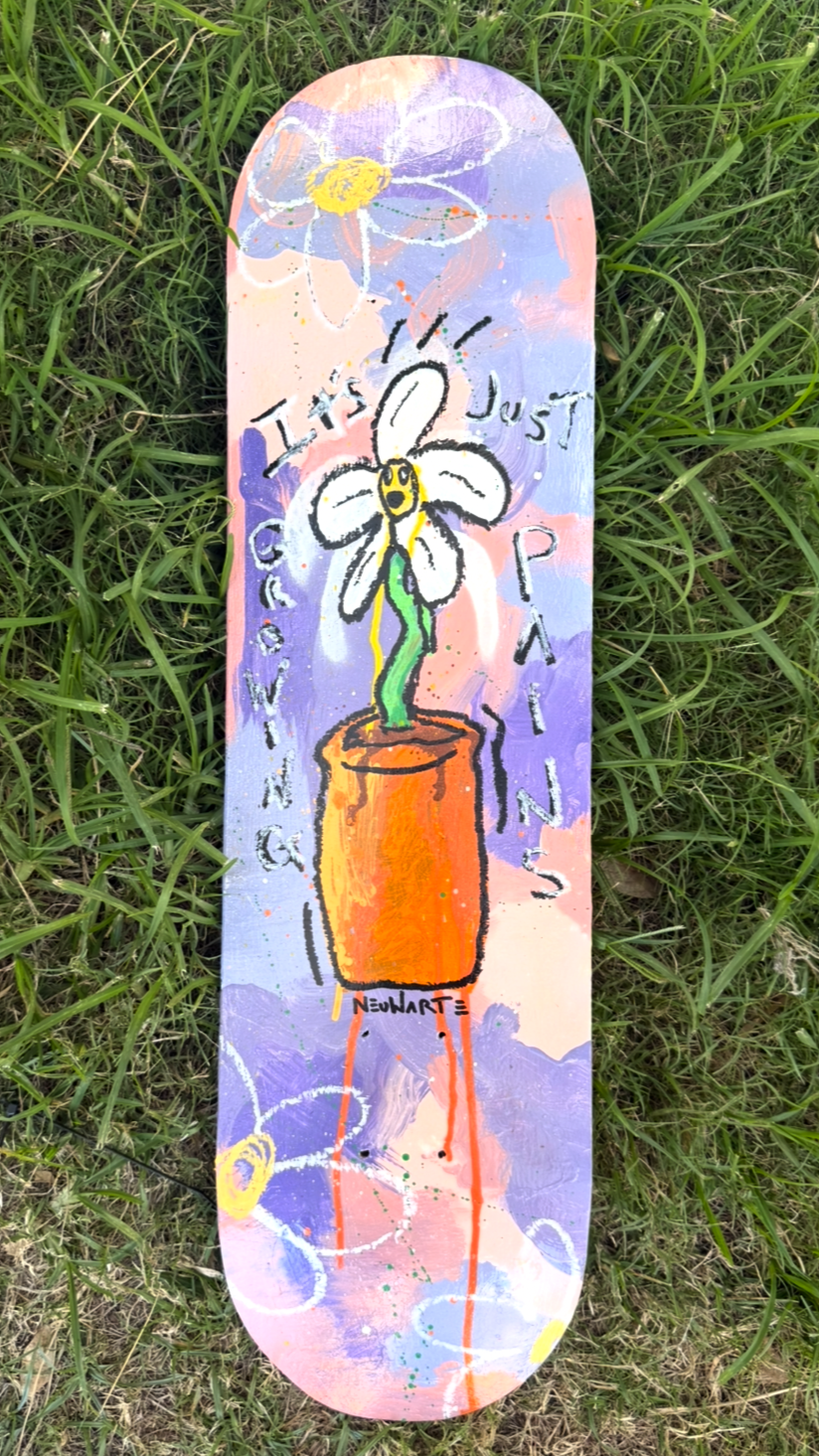 A colorful skateboard deck with a hand-painted design featuring a white flower with a yellow face in a pot, surrounded by abstract shapes and the words "It's just a quick painting." The background has pastel shades of purple, pink, and blue with cosm