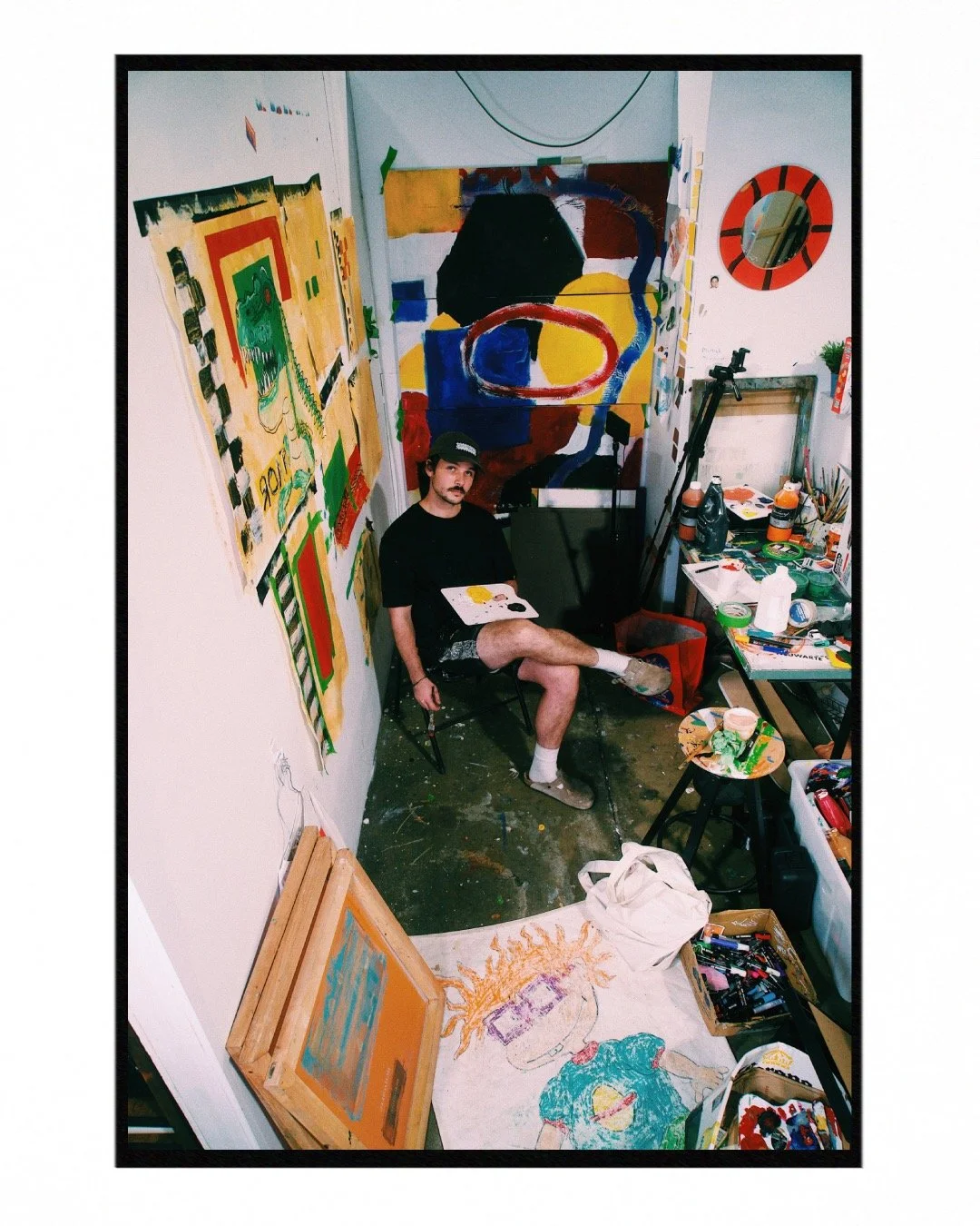 A young man in casual clothing and a baseball cap sitting in a small art studio, surrounded by colorful abstract paintings and art supplies. He has a small canvas on his lap and is seated on a black chair amidst vibrant paintings, brushes, and art materials.
