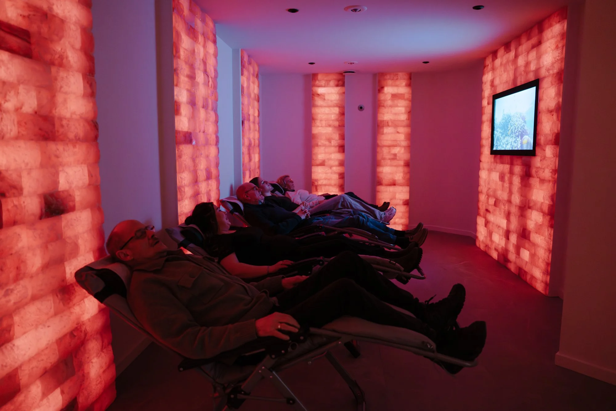Mensen liggen op ligstoelen in een zoutkamer met roze/oranje verlichte zoutwanden en een tv-scherm op de muur.