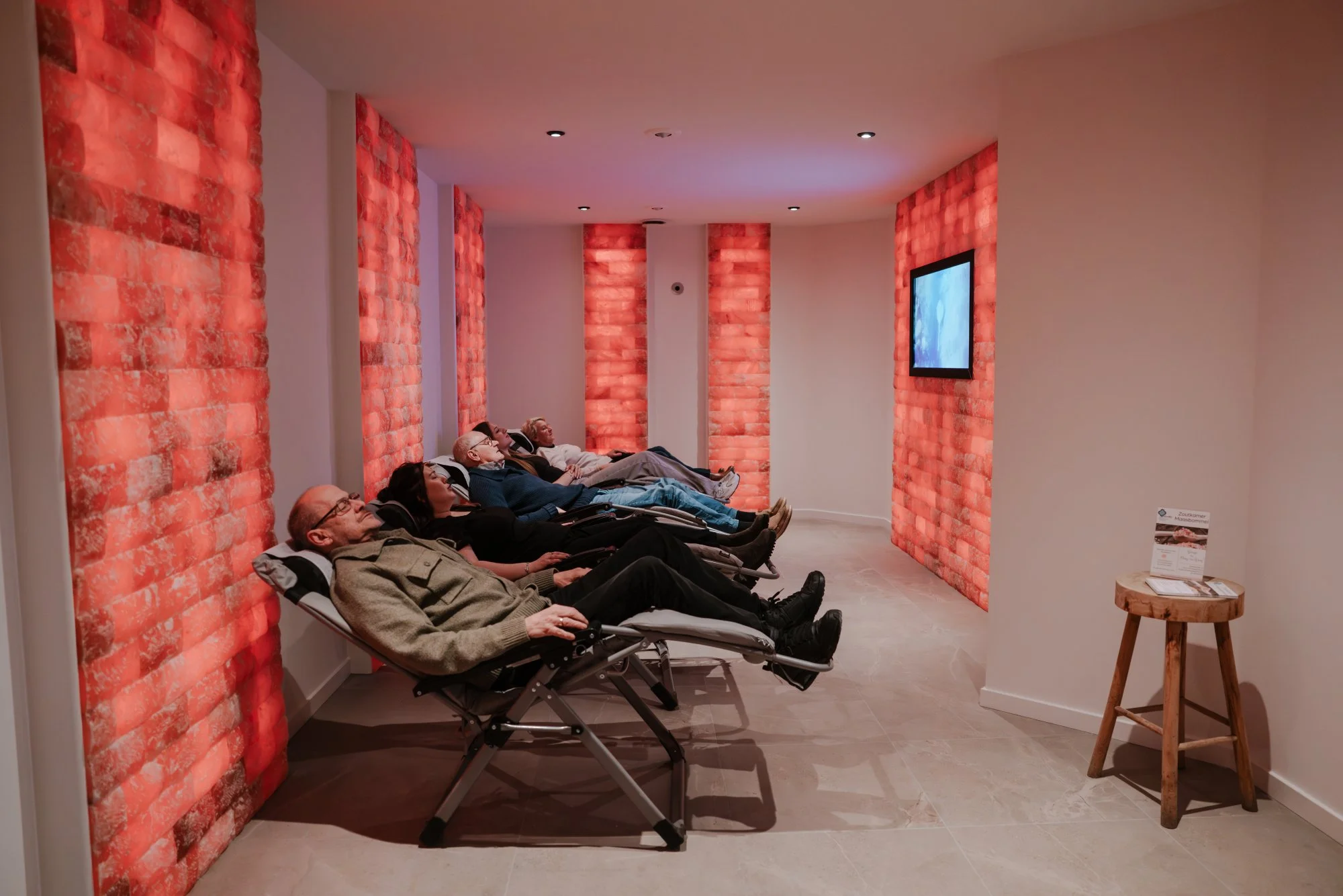 Vijf mensen liggen ontspannen in een zoutkamer, vlakbij een televisie met rode zoutstenen muur erachter.