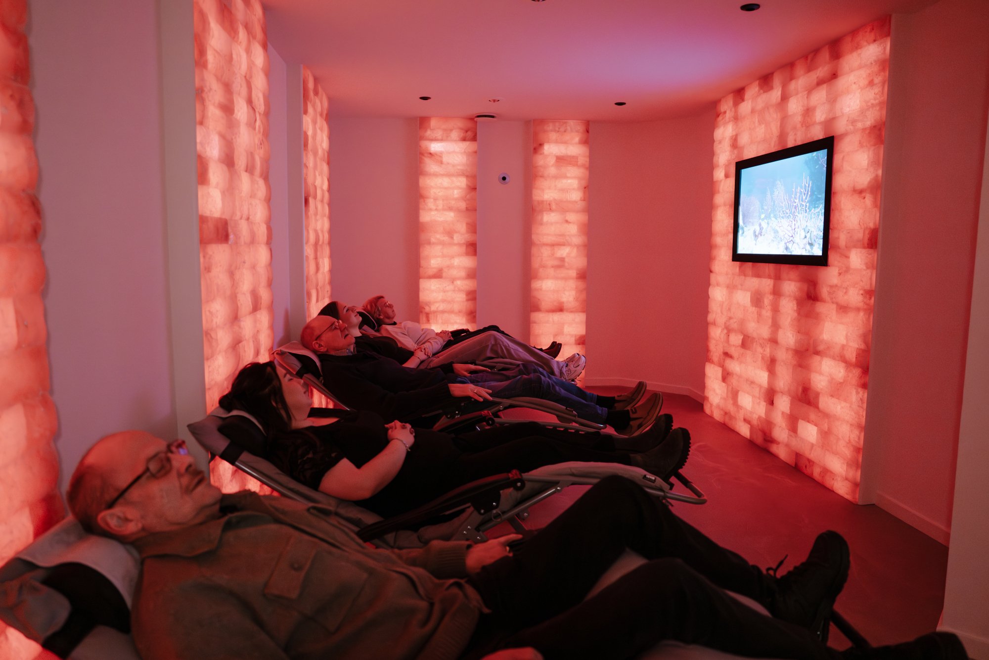 Vijf mensen liggen in een zoutkamer op ligstoelen en op een scherm worden relaxbeelden afgespeeld