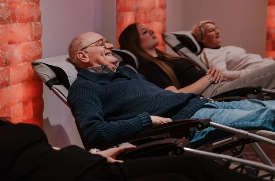 Drie mensen liggen op ligstoelen, ontspannen en lachend in een kamer met rode zoutstenen muur.