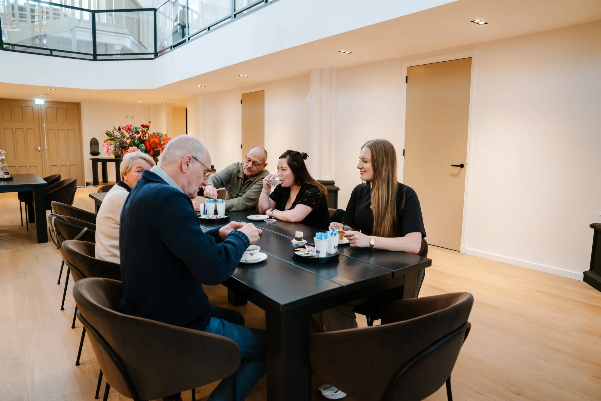 Vijf mensen zitten aan een zwarte tafel in de verbouwde kerk en genieten van koffie en snacks, met bloemen op de achtergrond.