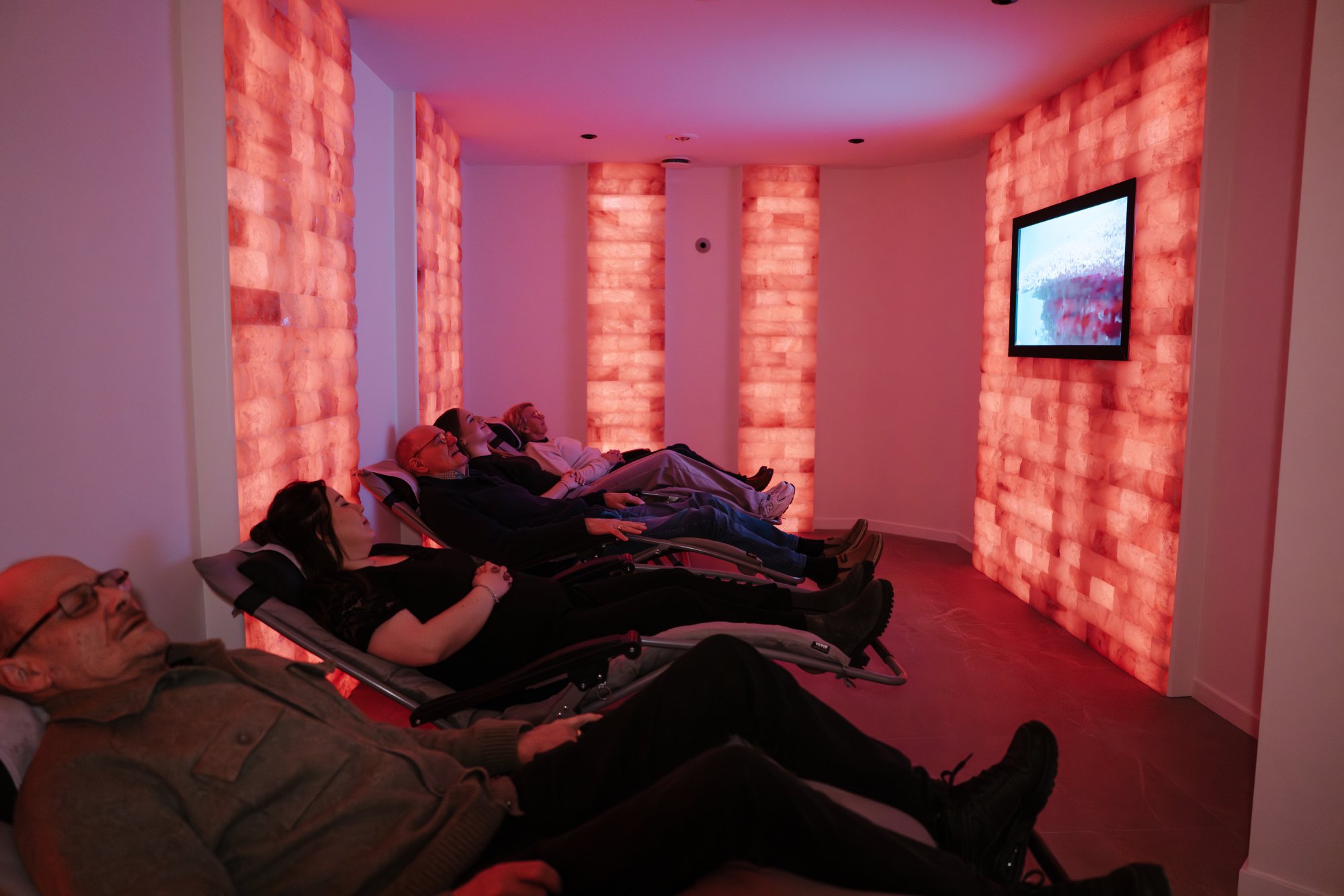 Mensen liggen op relaxstoelen in een zoutkamer met roze/oranje verlichte zoutstenen muren, kijken naar een televisie.