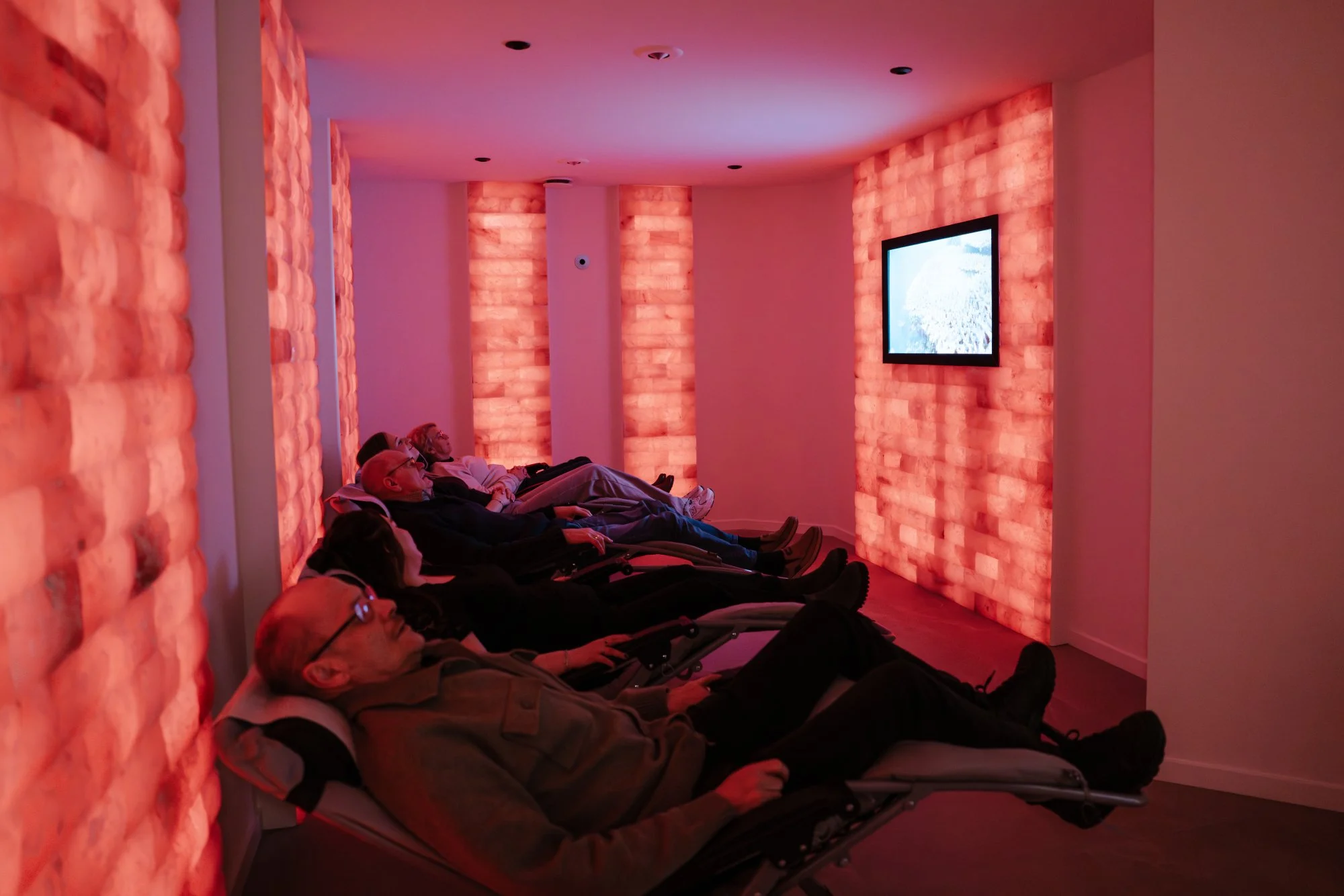 Mensen liggen in een zoutkamer met sfeervolle rode verlichting en kijken naar een scherm aan de muur.