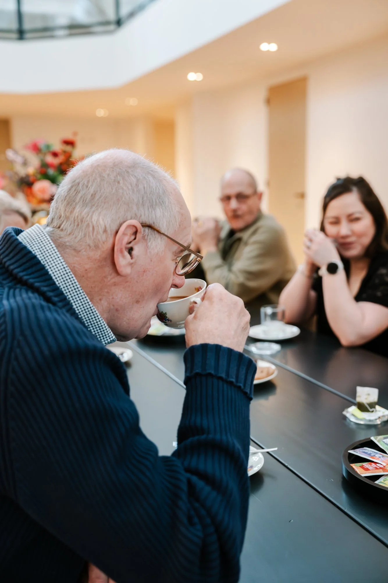 Een bezoeker drinkt thee in een groep mensen die aan een tafel zitten, op een warme, lichte binnenlocatie.