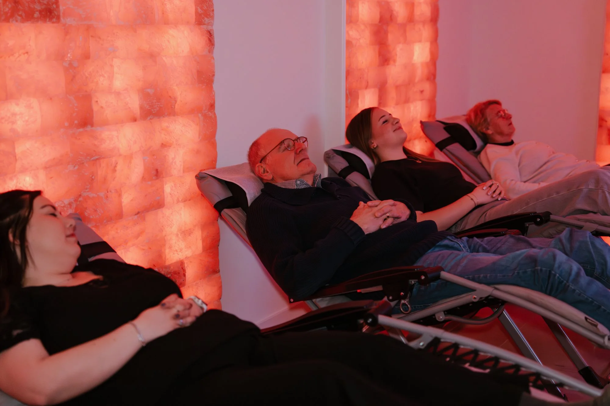 Vier mensen liggen op ligstoelen in een zoutkamere met een oranje zoutmuur op de achtergrond, ze lijken te ontspannen.