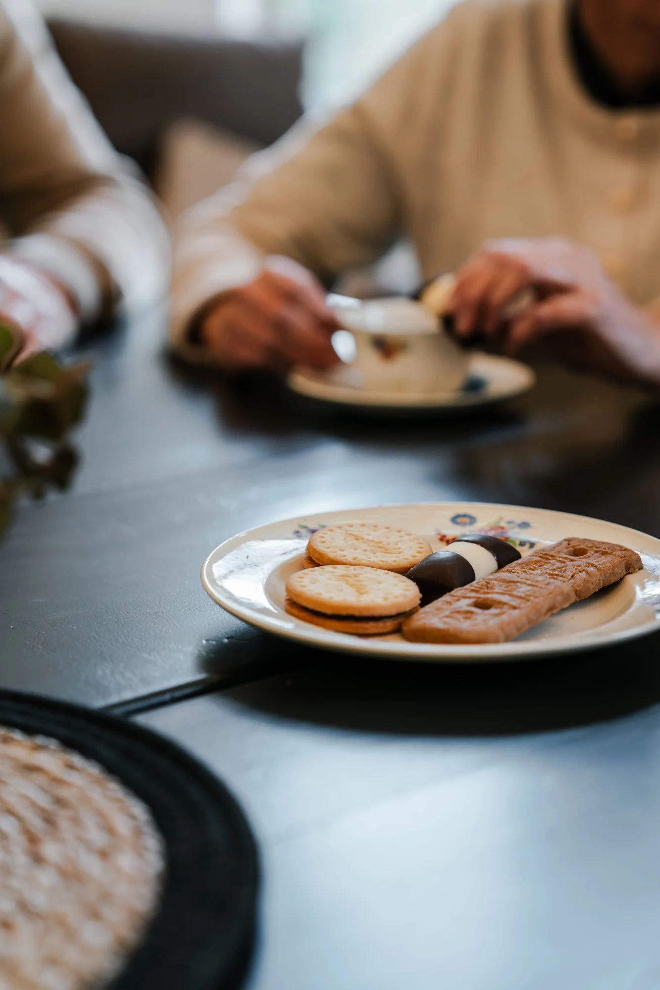 Een wit bord met een stuk chocolade, en een koekje op een donkere tafel, met twee mensen op de achtergrond die aan tafel zitten.