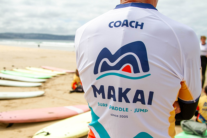 MAKAI coach wearing branded clothing beside surfboards on the beach.