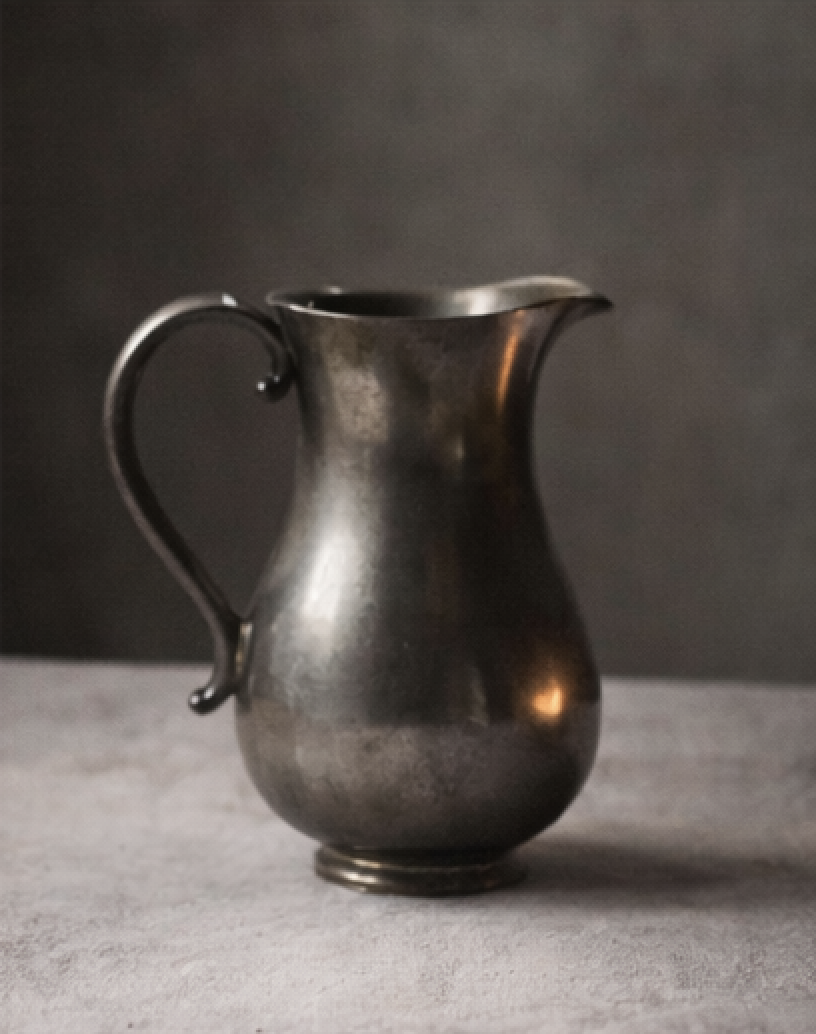 A metallic pitcher with a curved handle and a wide spout, set against a dark background.
