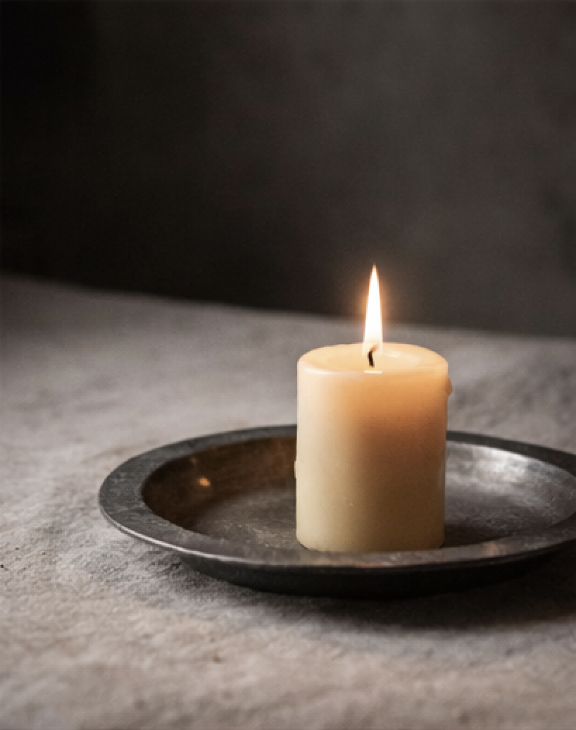 A lit white pillar candle on a black metal plate, placed on a textured gray surface with dark background.