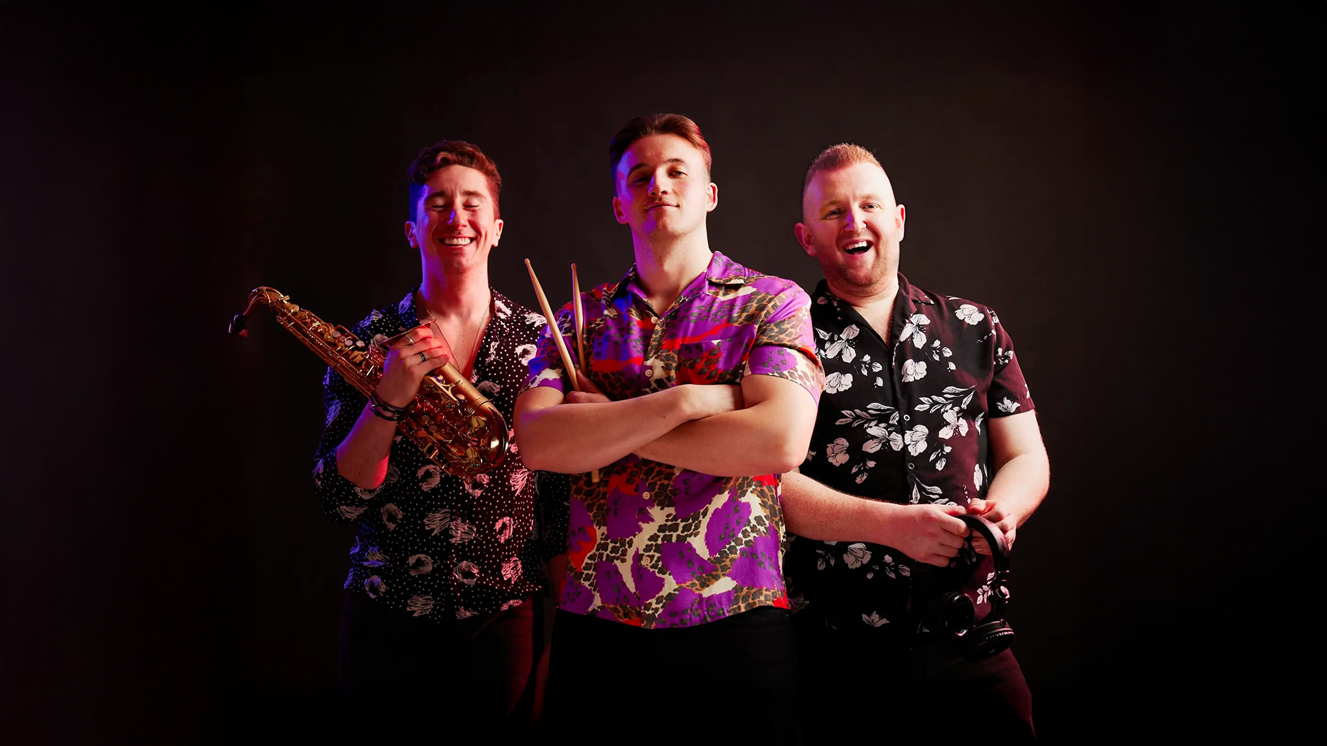 Three men wearing colorful floral shirts, standing against a black background. The man on the left holds a saxophone, the man in the center has drumsticks crossed over his chest, and the man on the right holds a camera.