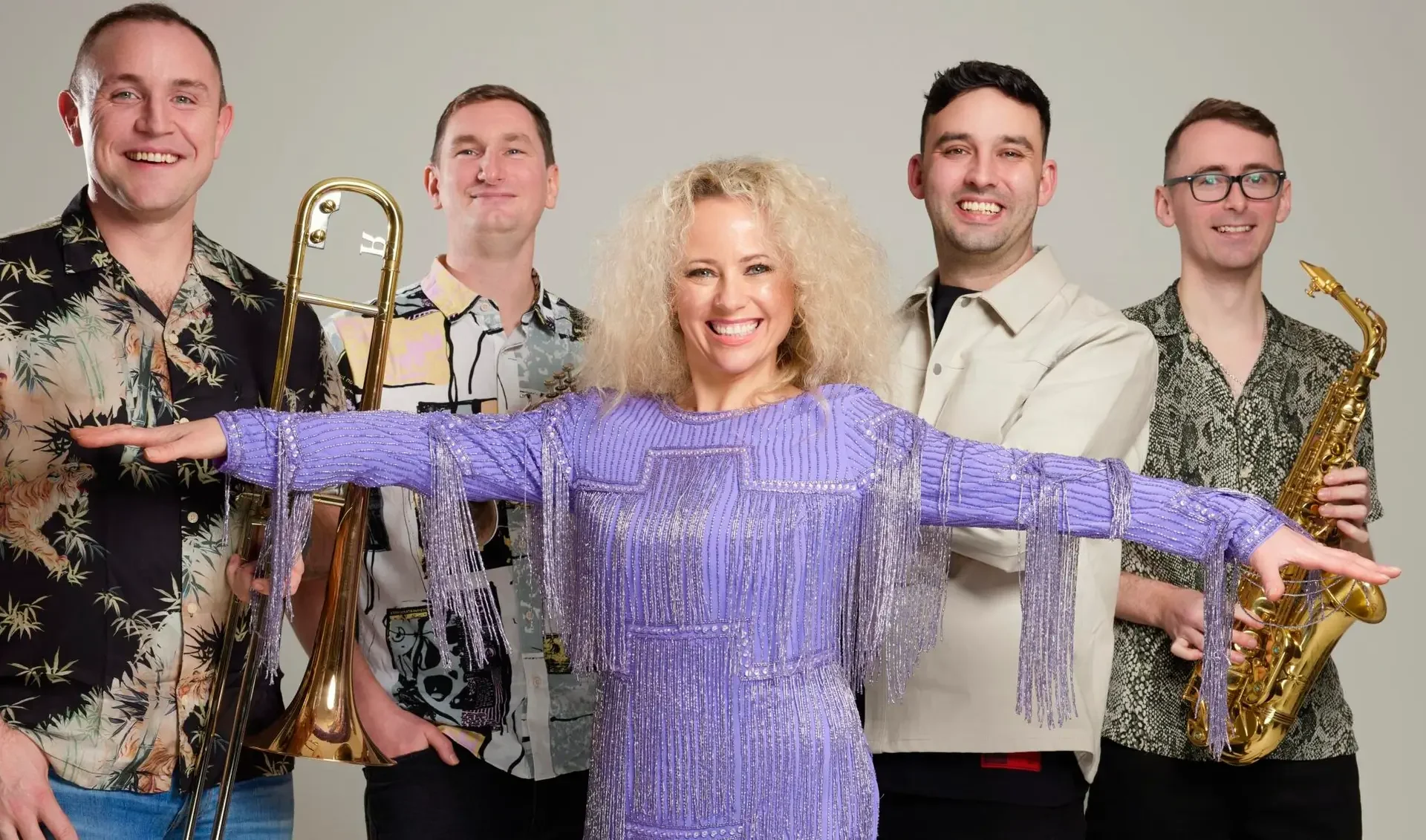 A group of five musicians, four men and one woman, standing with musical instruments against a light background, woman in purple dress with arms outstretched.