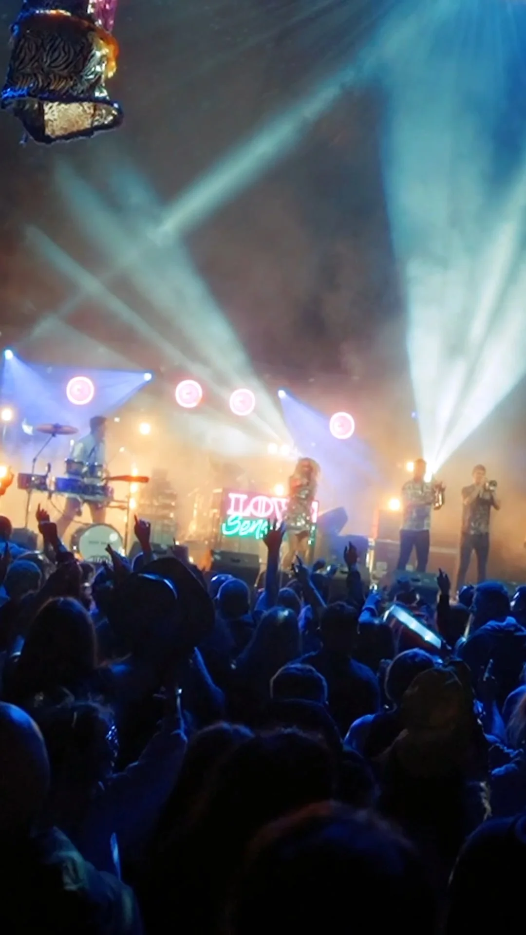 A live concert with a band performing on stage, illuminated by colorful lights and smoke, with an audience waving their hands.