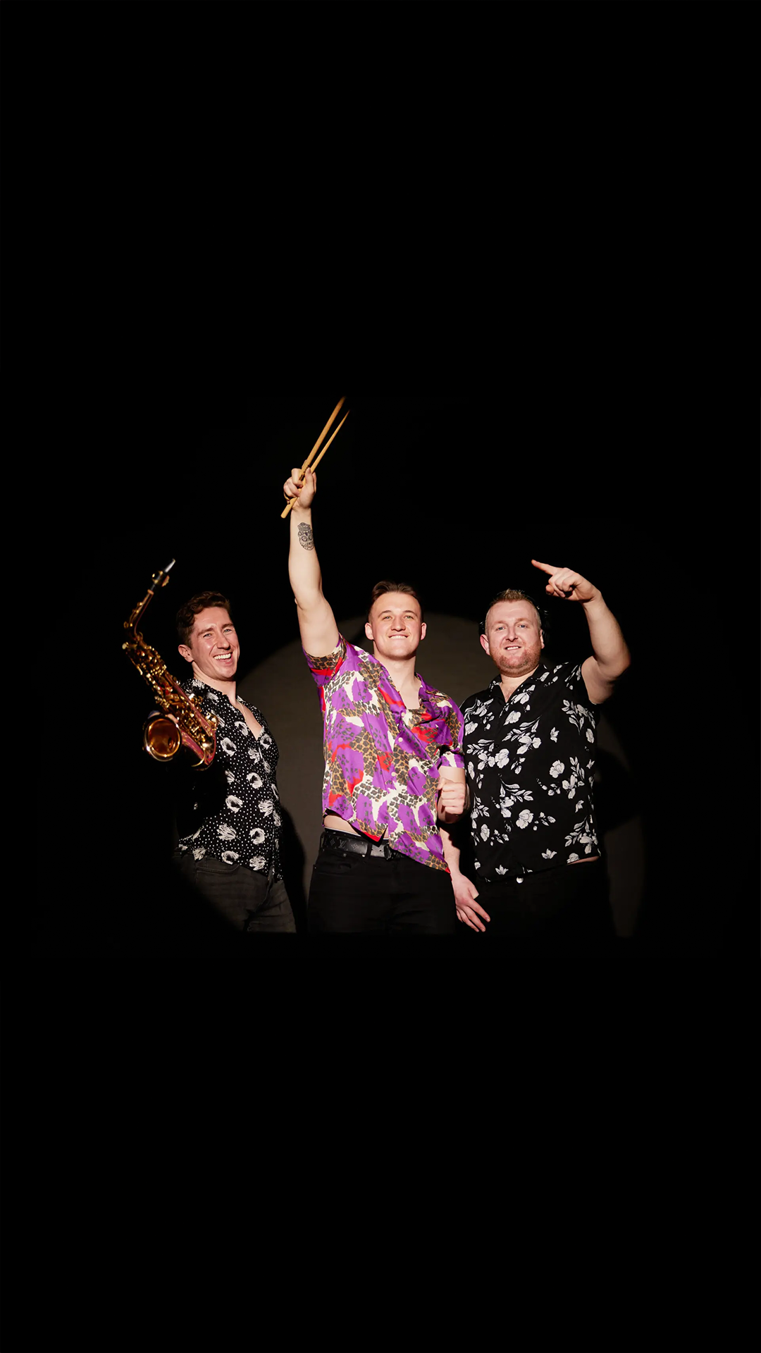 Three men dressed in colorful, patterned shirts smiling on stage. One man is holding a saxophone, the second is raising a pair of drumsticks, and the third is pointing with his finger. The background is black.