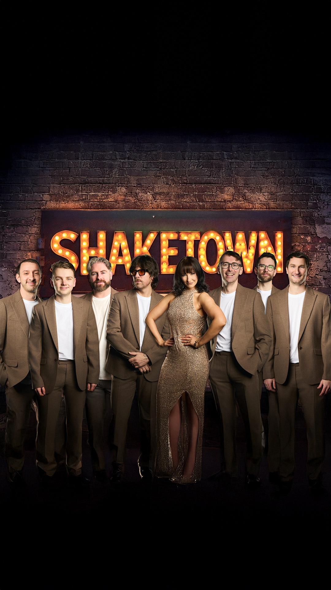 A group of seven men and one woman dressed in formal attire, standing in front of a brick wall with illuminated sign reading 'SHAKETOWN'.