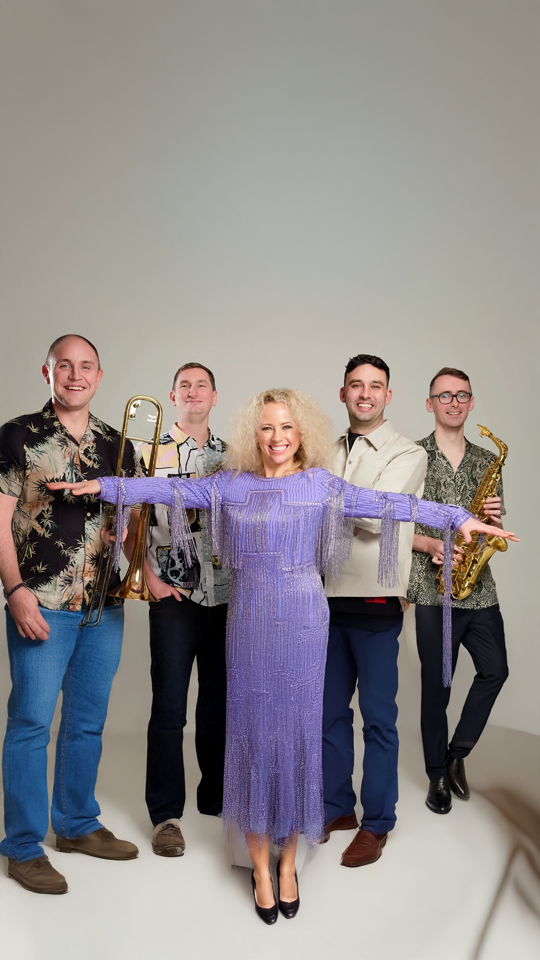 A woman in a purple dress stands in front of a group of five men, holding out her arms as if conducting an orchestra, with music instruments in their hands, against a plain background.
