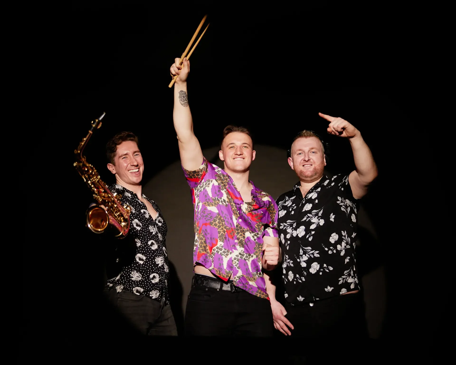 Three men in colorful floral shirts celebrating on stage, with one holding chopsticks and a saxophone, and the man on the right pointing upward.