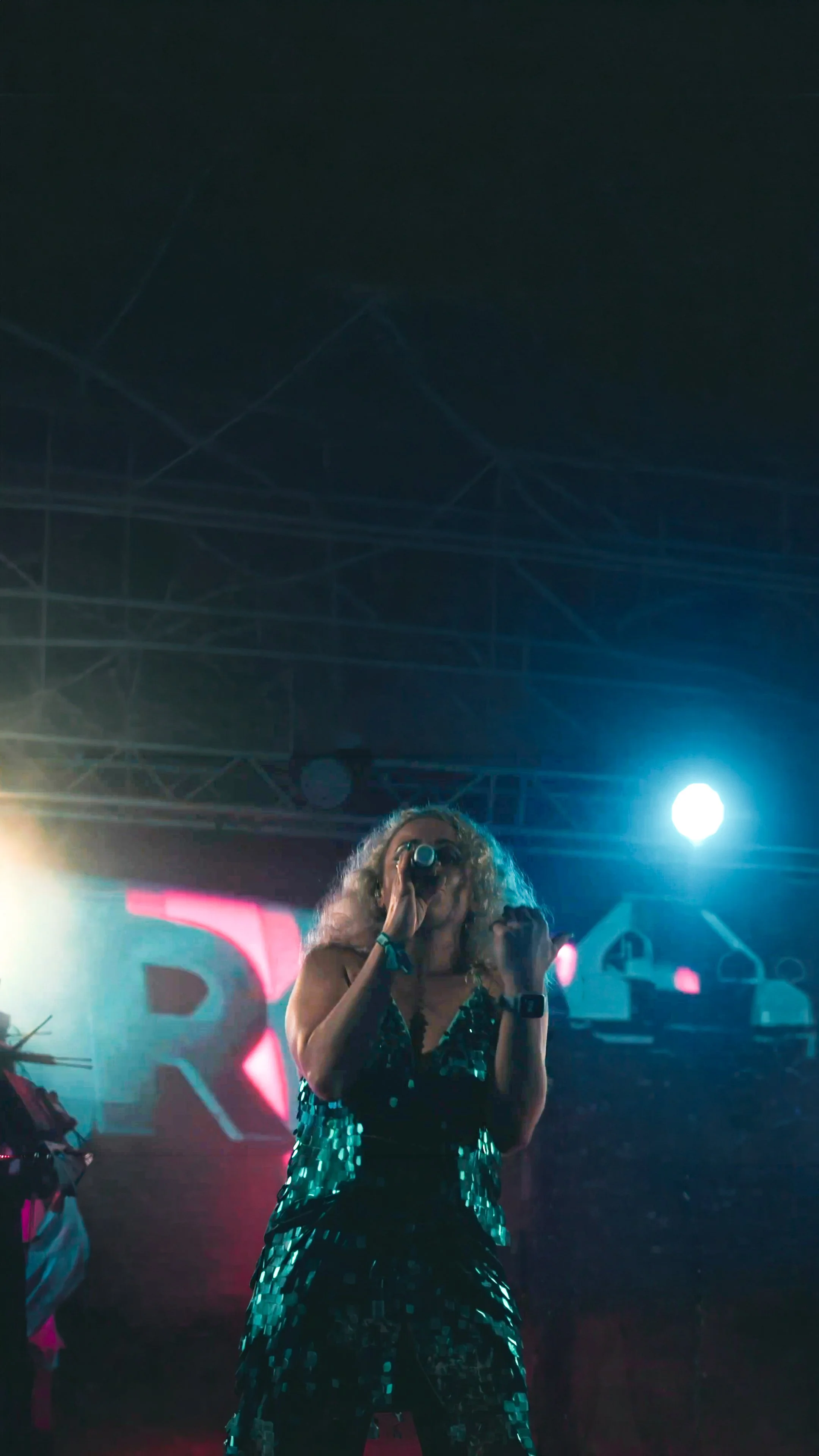 A woman with curly blonde hair singing into a microphone on stage with colorful lighting and a large pink and white backdrop.