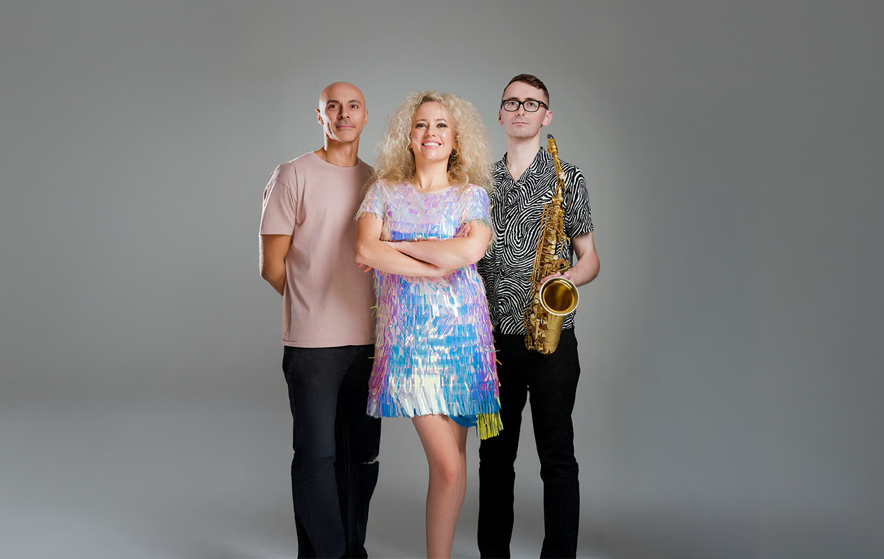 Three musicians standing together: a woman in a colorful dress smiling with arms crossed, a man in a striped shirt holding a saxophone, and another man in a beige shirt. Gray background.