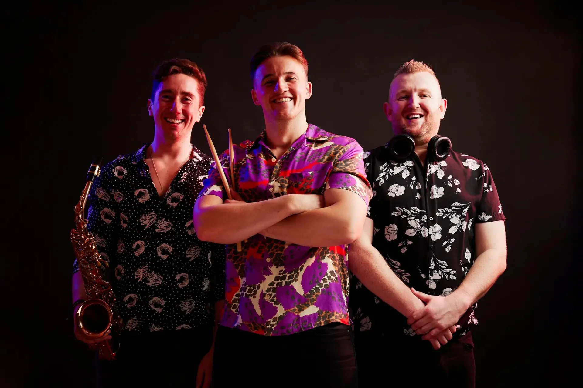 Three young men standing together against a black background, smiling, wearing colorful floral shirts, with one holding a saxophone and one with headphones around his neck.