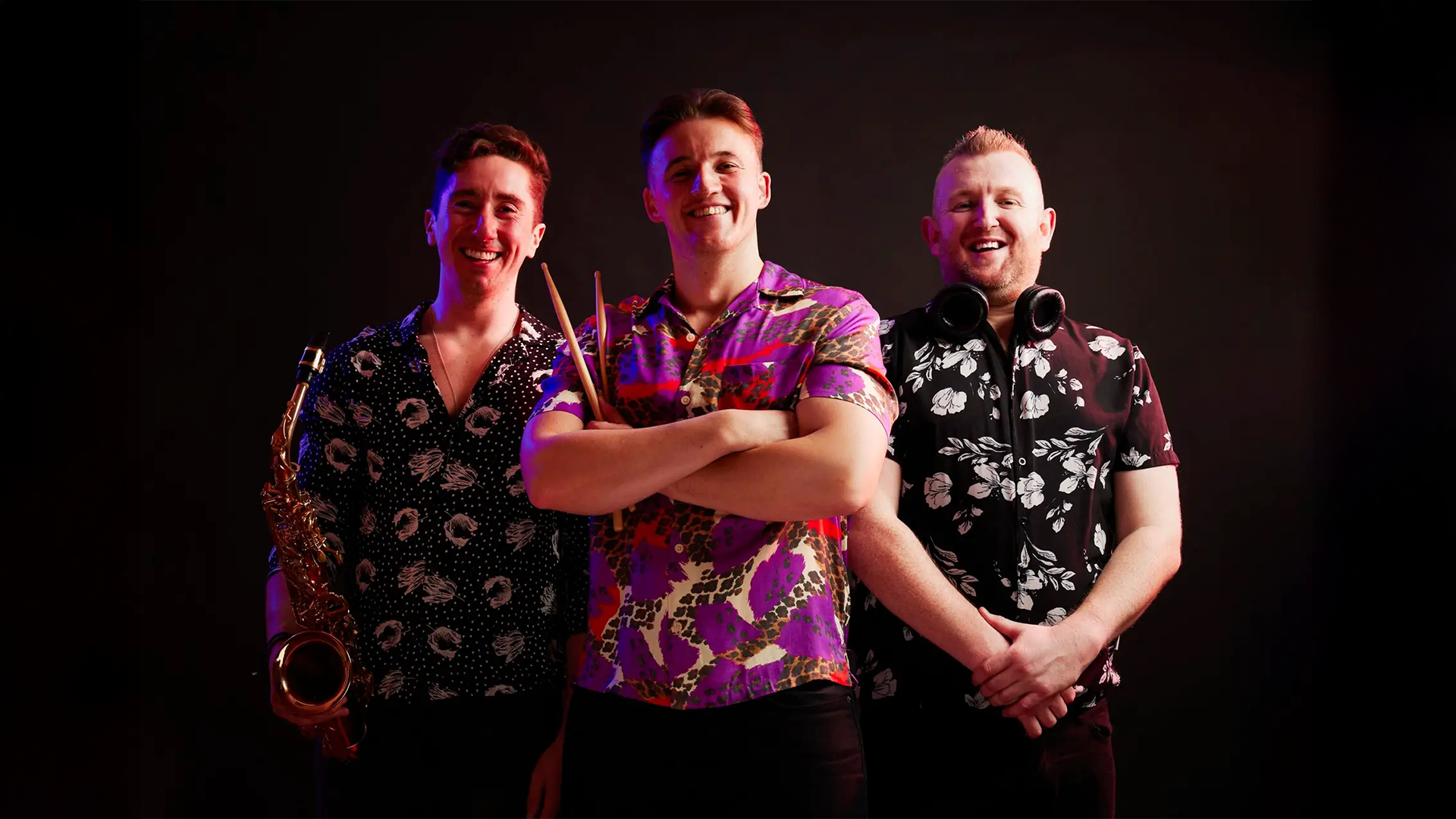 Three men standing against a black background, smiling at the camera. The man on the left holds a saxophone, the man in the middle crosses his arms and holds drumsticks, and the man on the right has headphones around his neck. All are wearing colorful, patterned short-sleeve shirts.