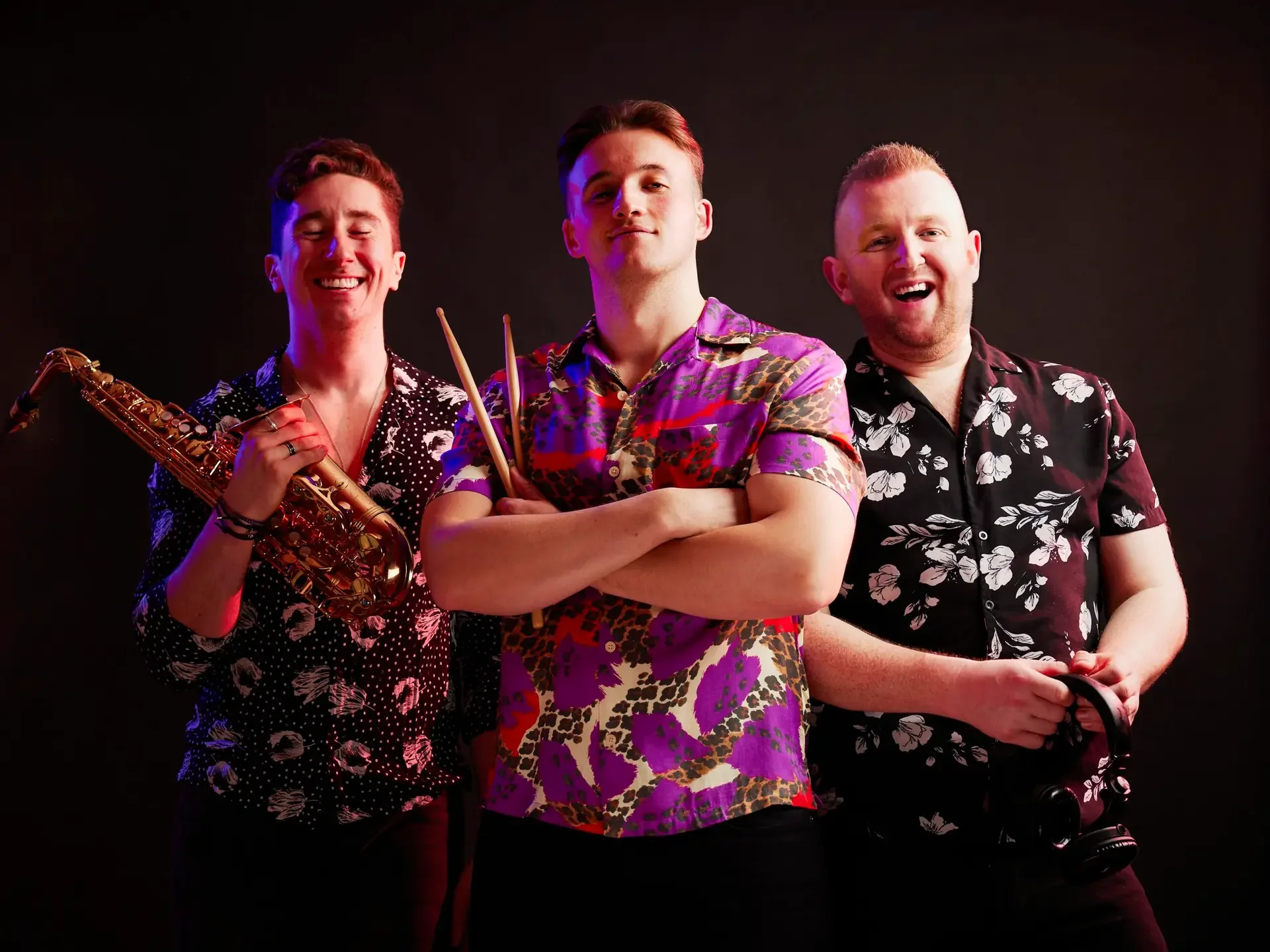Three young men dressed in colorful floral shirts standing in front of a dark background, with one holding a saxophone, another with arms crossed holding drumsticks, and the third holding headphones, smiling and looking at the camera.