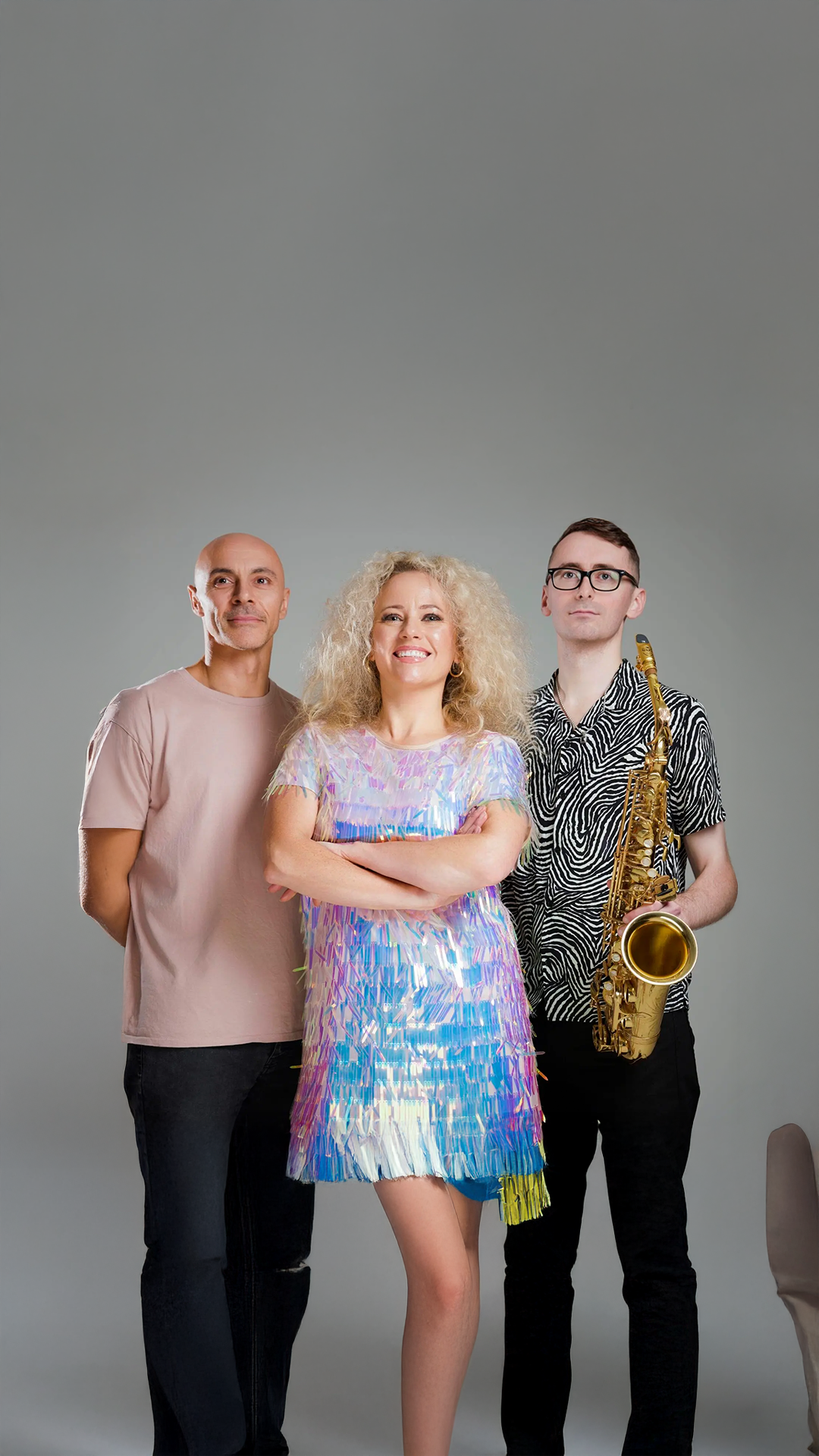 Group of three people, two men and one woman, posing together. The woman in the center is smiling, has blonde curly hair, and is wearing a colorful, shiny dress. The man on the right has glasses, short dark hair, and is holding a saxophone. The man on the left is bald, wearing a plain beige T-shirt.
