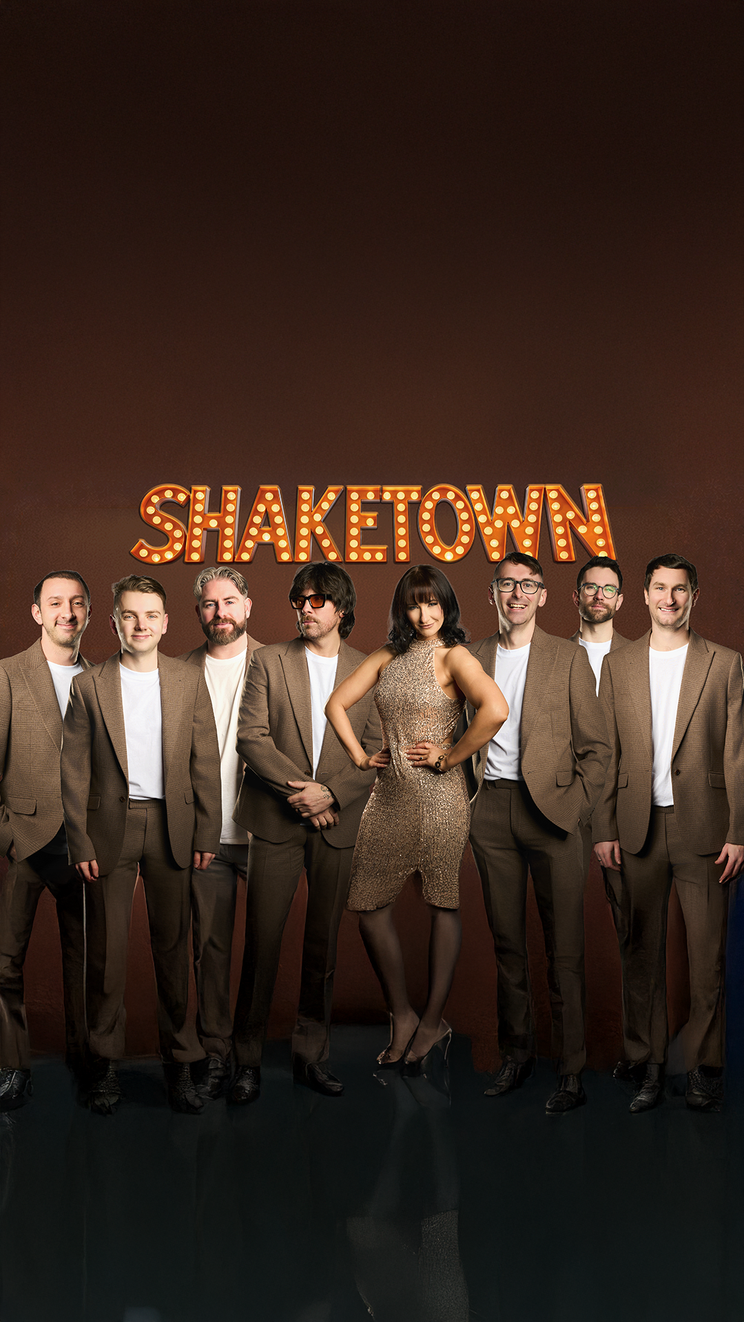 A group of seven people standing in front of a dark brown background with a sign that says "SHAKETOWN" in marquee lights. The woman in the center is wearing a gold dress, and the men are dressed in brown suits with white shirts.