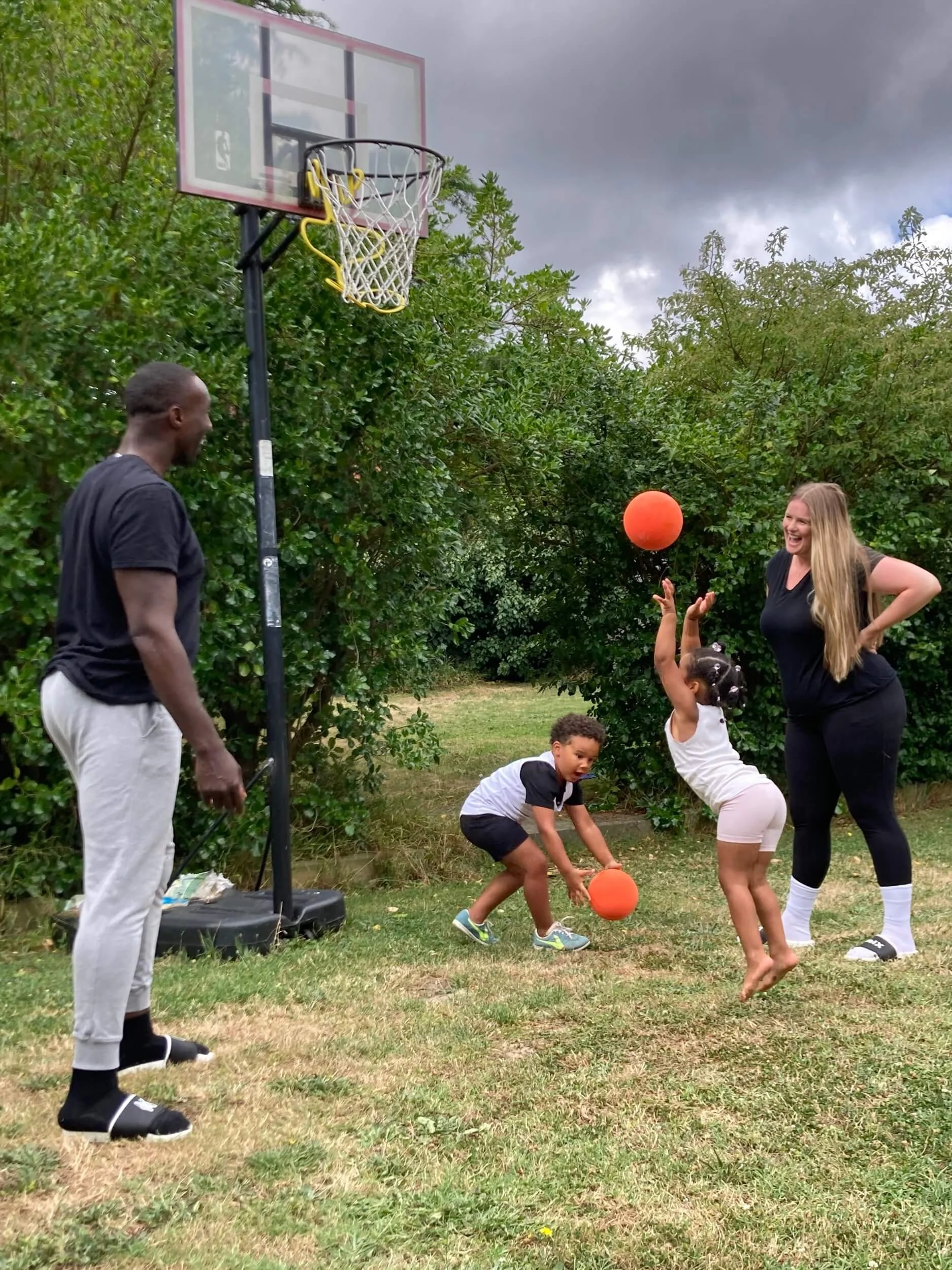 A family playing basketball outdoors on a cloudy day. A young girl is jumping to shoot a basketball, while a boy is holding another basketball, and an adult woman is standing nearby smiling. An adult man is also present near the basketball hoop.