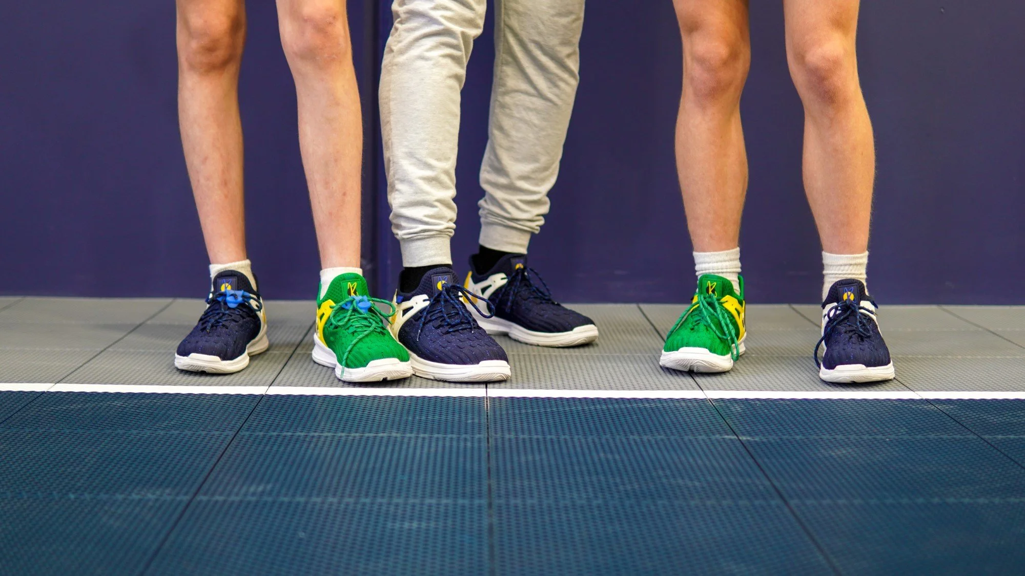 Photo showing the legs and feet of three people standing side by side on a gym floor, wearing athletic shoes in blue, green, and black, with a dark blue wall in the background.