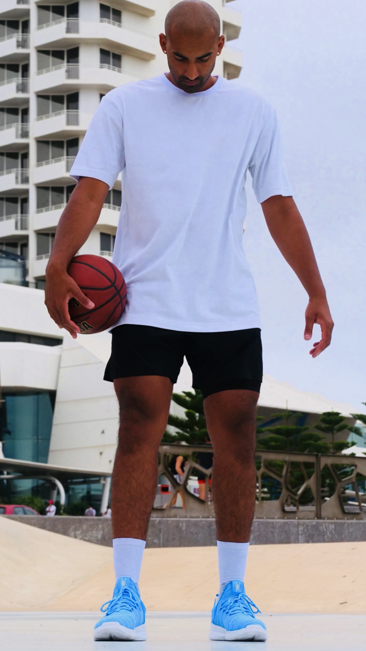 Young man in white sports shirt and black shorts holding a basketball, standing outdoors with modern buildings and trees in the background.