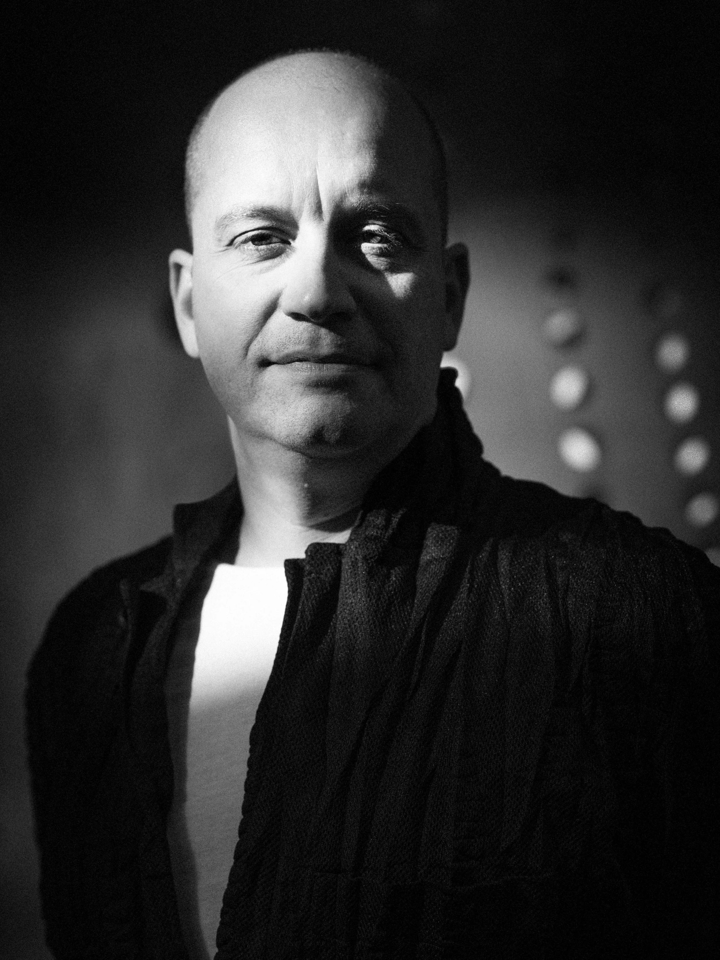 Black and white portrait of a bald man with a slight smile, wearing a dark textured jacket and a white shirt, with a blurred background featuring circular lights.