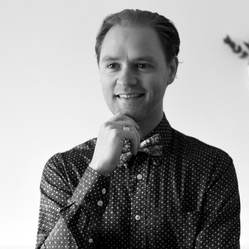 A young man with slicked-back hair, smiling, wearing a patterned shirt and bow tie, posed with his hand on his chin in a reflective manner.