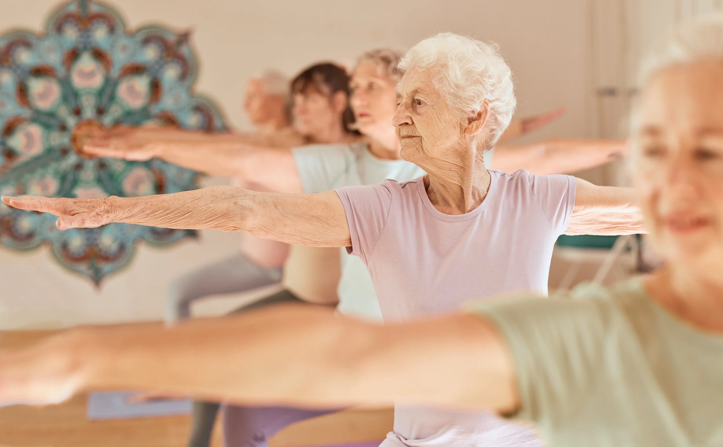 Senior women participating in a seated yoga or stretching class in a bright studio, extending their arms out to the sides.