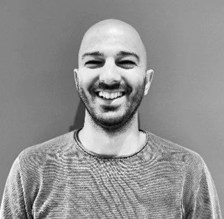 A smiling man with a bald head and beard, wearing a patterned shirt, standing in front of a plain wall.