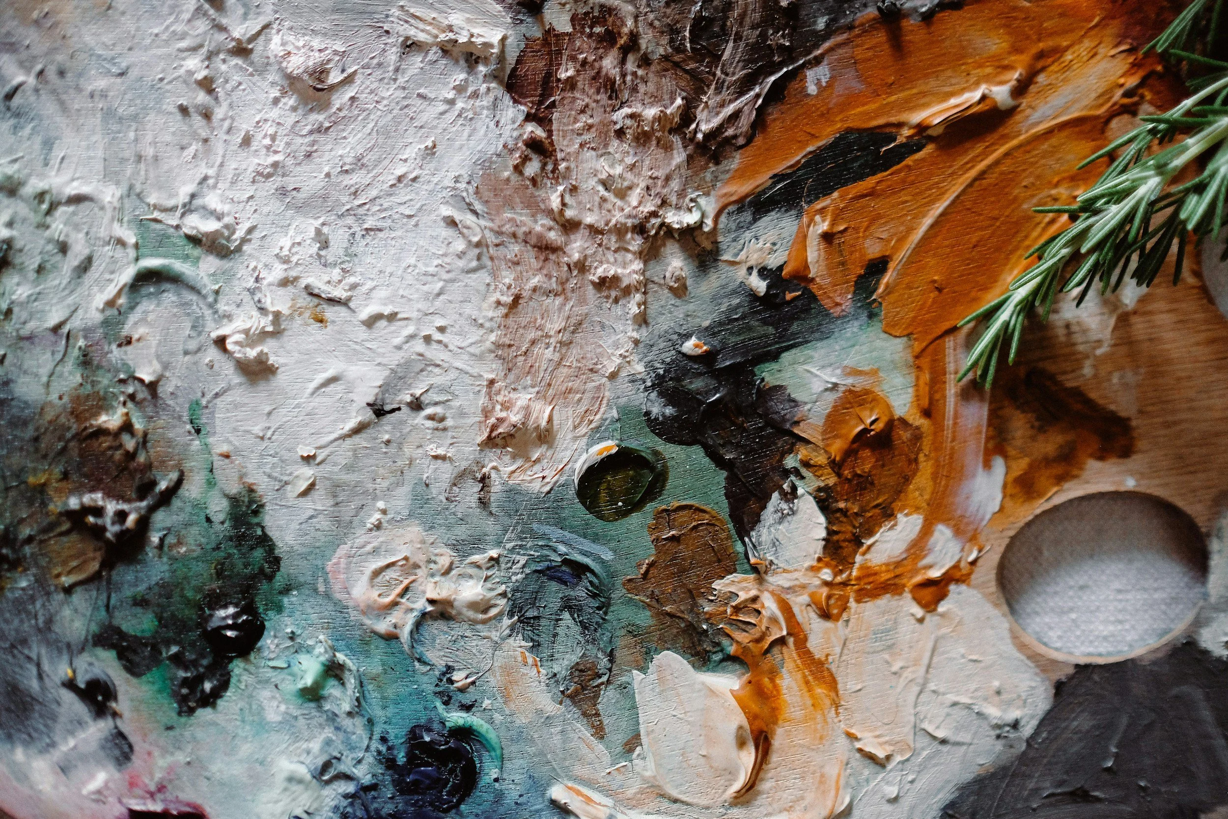 Close-up of an artist's palette with thick, textured oil paint in various colors including white, black, orange, and green, with a sprig of pine in the upper right corner.