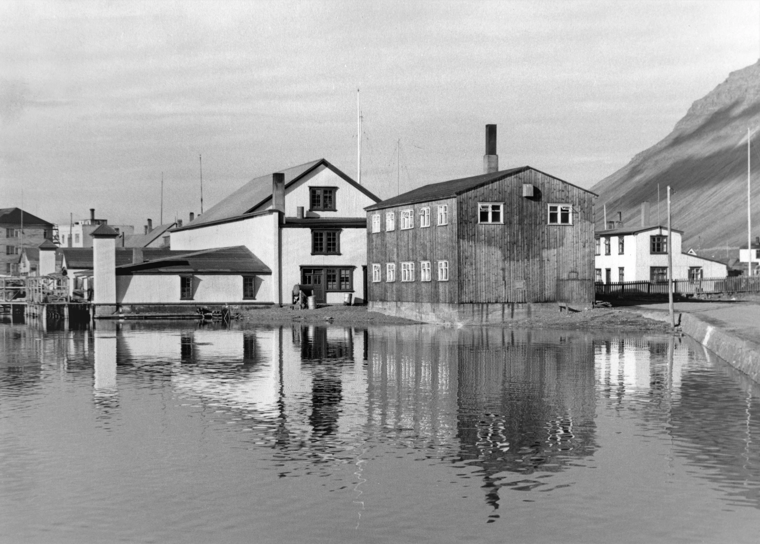 Black and white photograph of Edinborgarhúsið and surrounding houses, near Pollurinn, with reflections, mountain in the background, and clear sky.