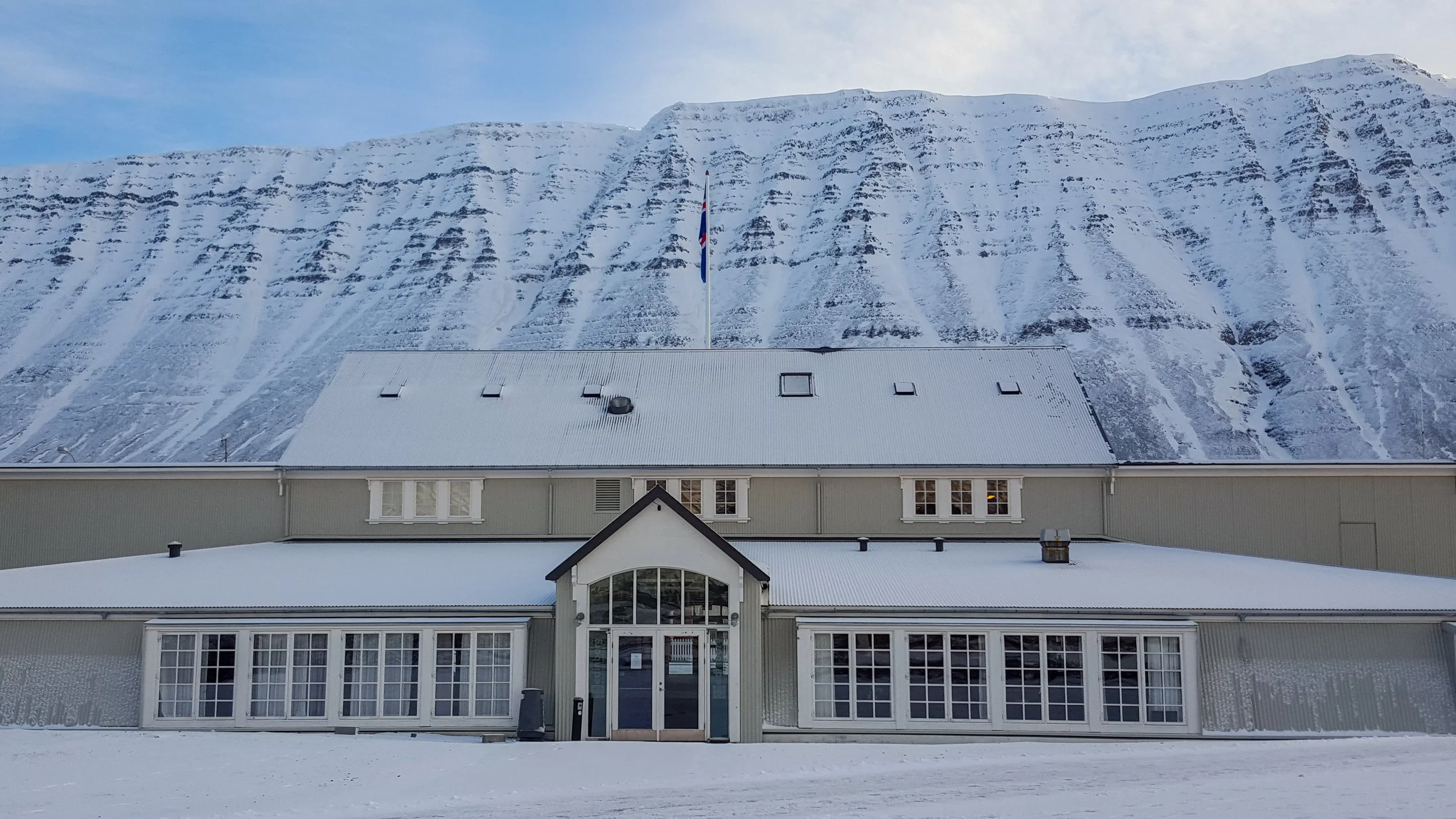 Edinborgarhúsið seen from Pollgataa. A large building with a glass entrance, beige walls, and a snow-covered roof, set against snow-covered mountains in the background.